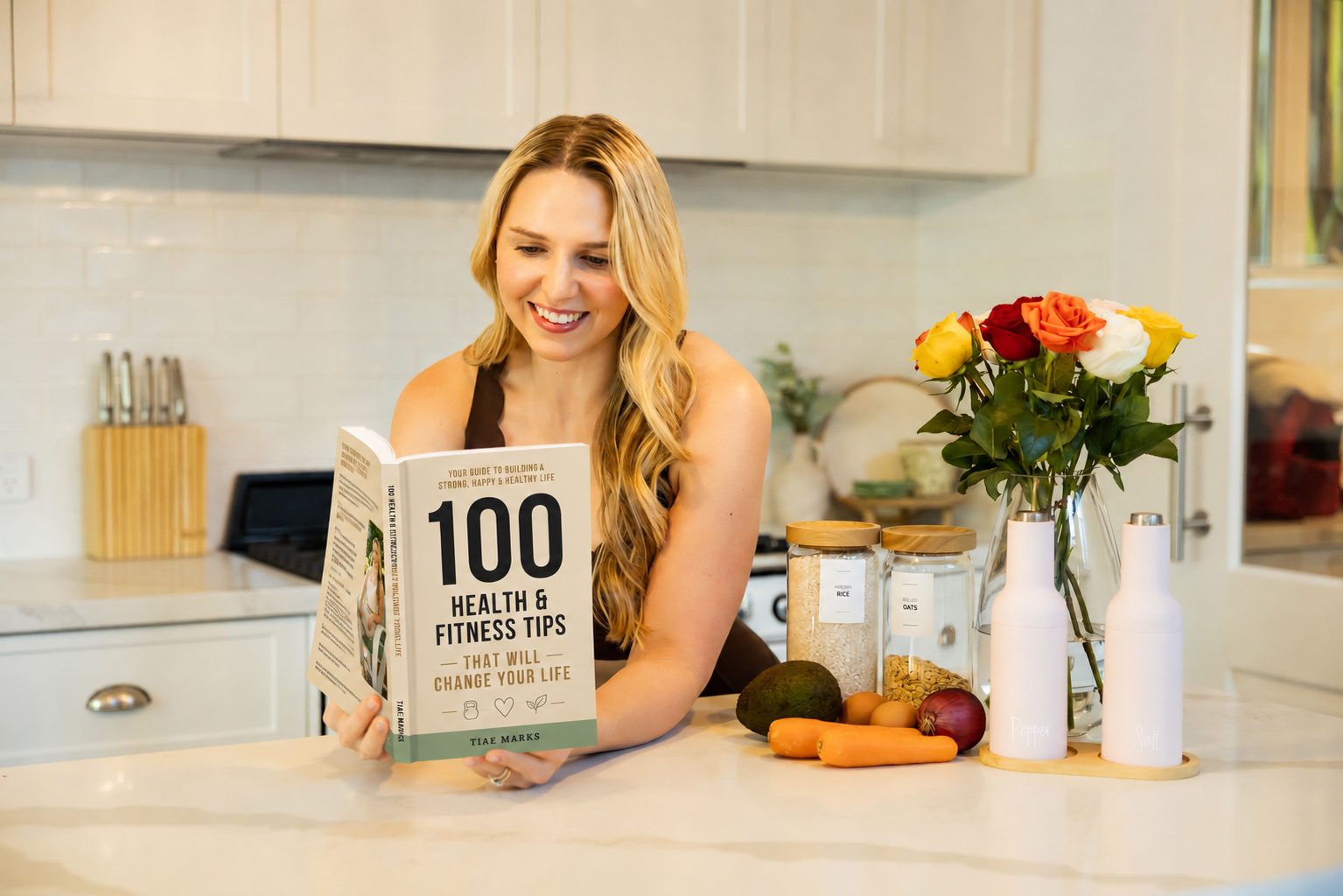 A woman with long blonde hair smiling and holding a book titled '100 Health & Fitness Tips That Will Change Your Life' in a bright kitchen. On the counter in front of her are vegetables, jars labeled 'Rice' and 'Oats,' and a vase with colorful roses.