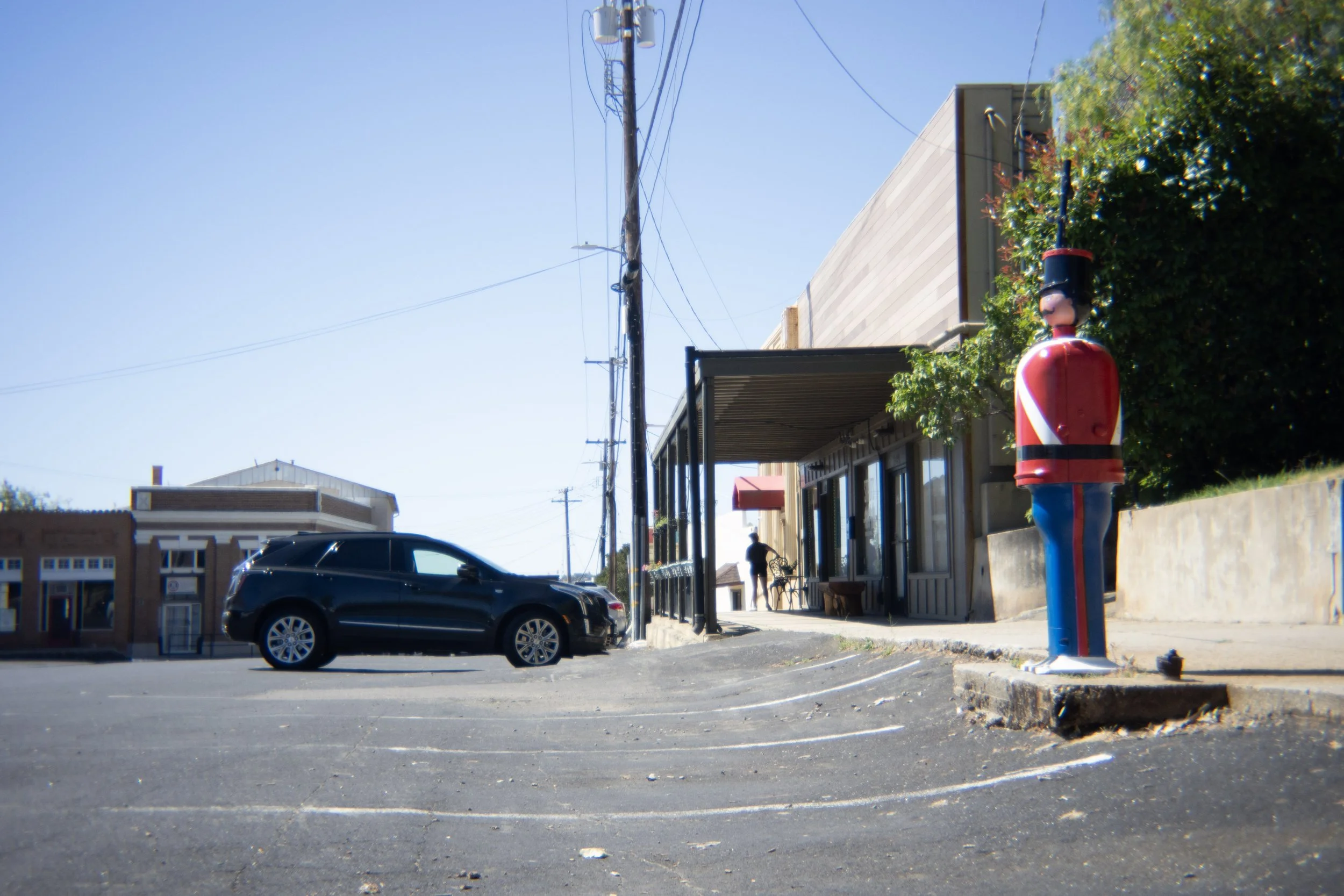A sunny street scene featuring a parking lot with some black cars, a building with a side porch, a person walking and a large decorative nutcracker figure near a green bush.