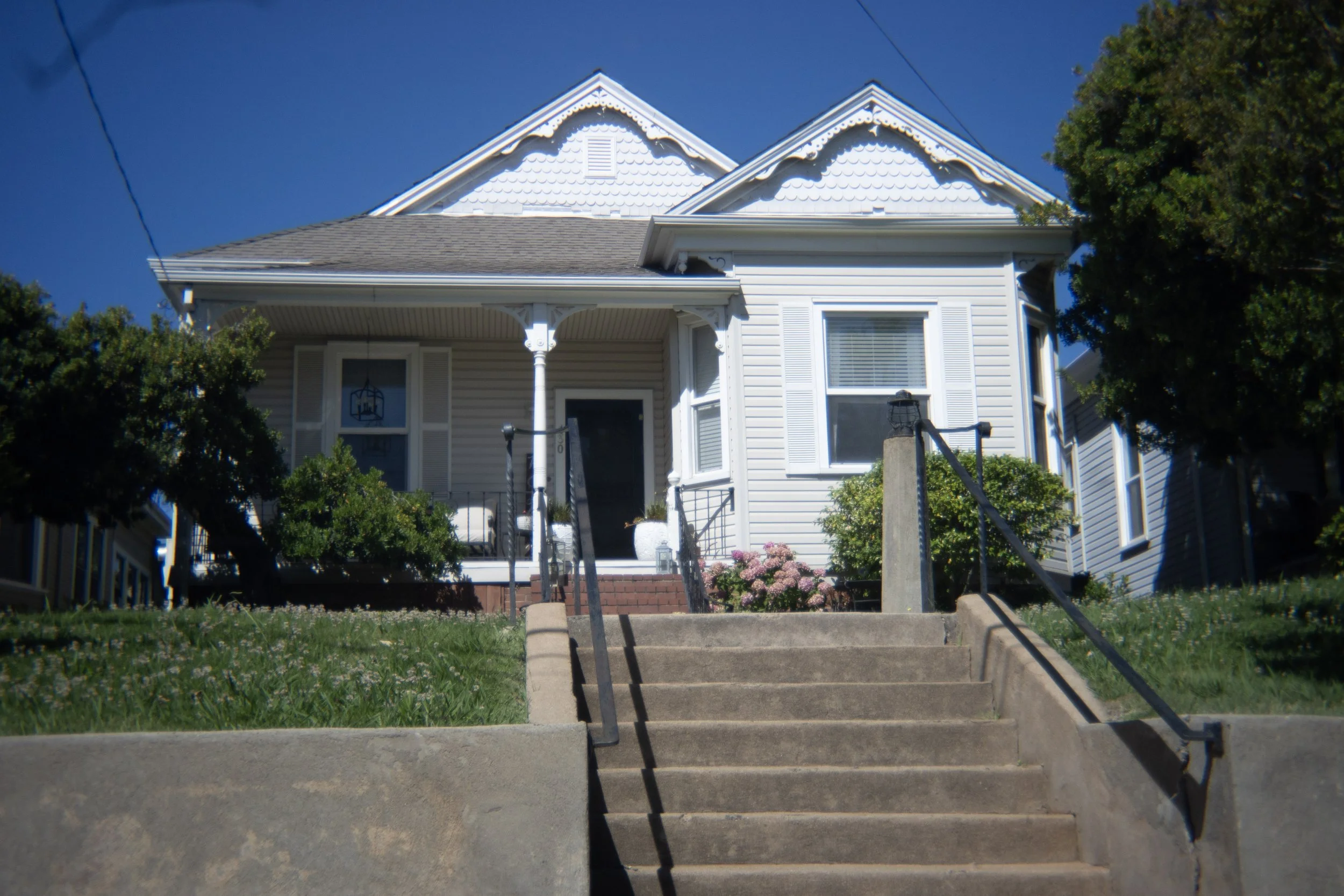 Front porch of a white, two-story house with decorative trim, shutters, and a small front garden with pink flowers. Stone stairs lead up to the porch, with metal railings on both sides.