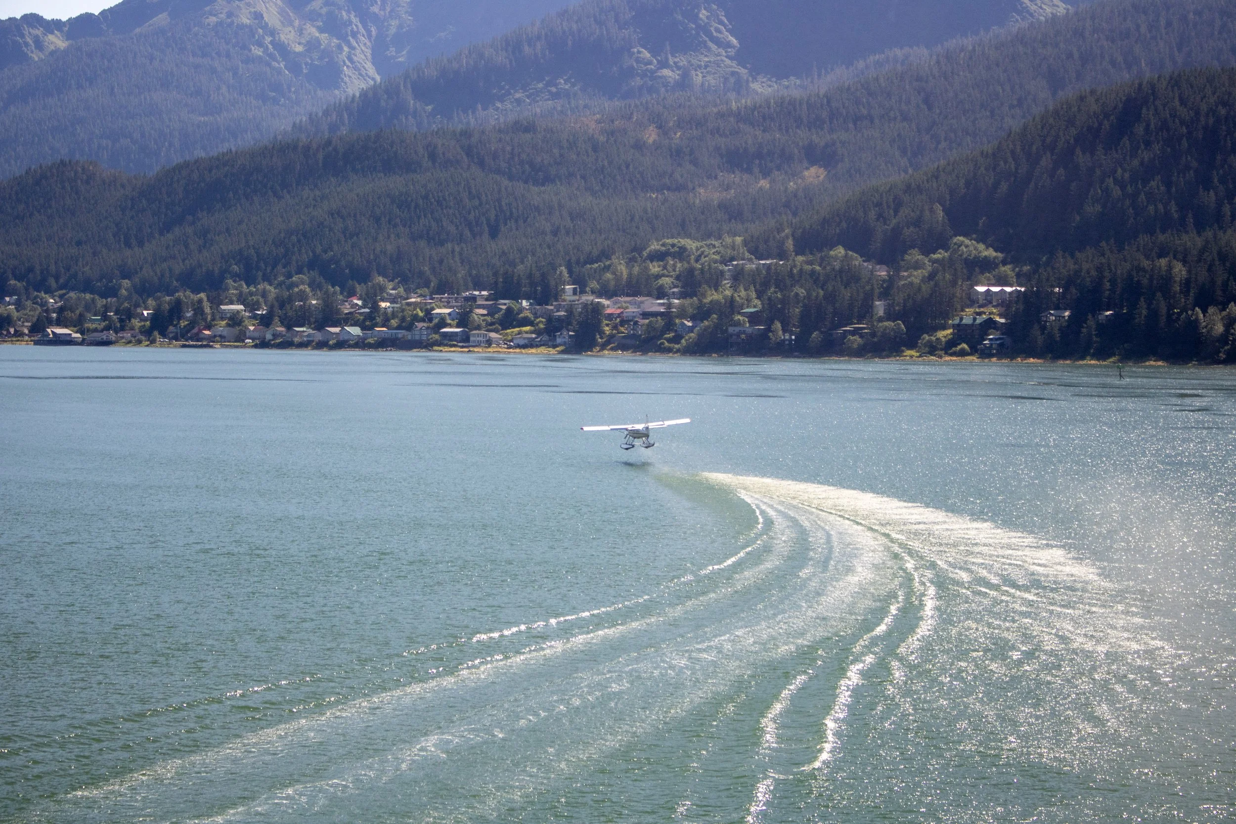 A small seaplane flying low over a body of water with a small wake behind it, with a town and forested mountains in the background.