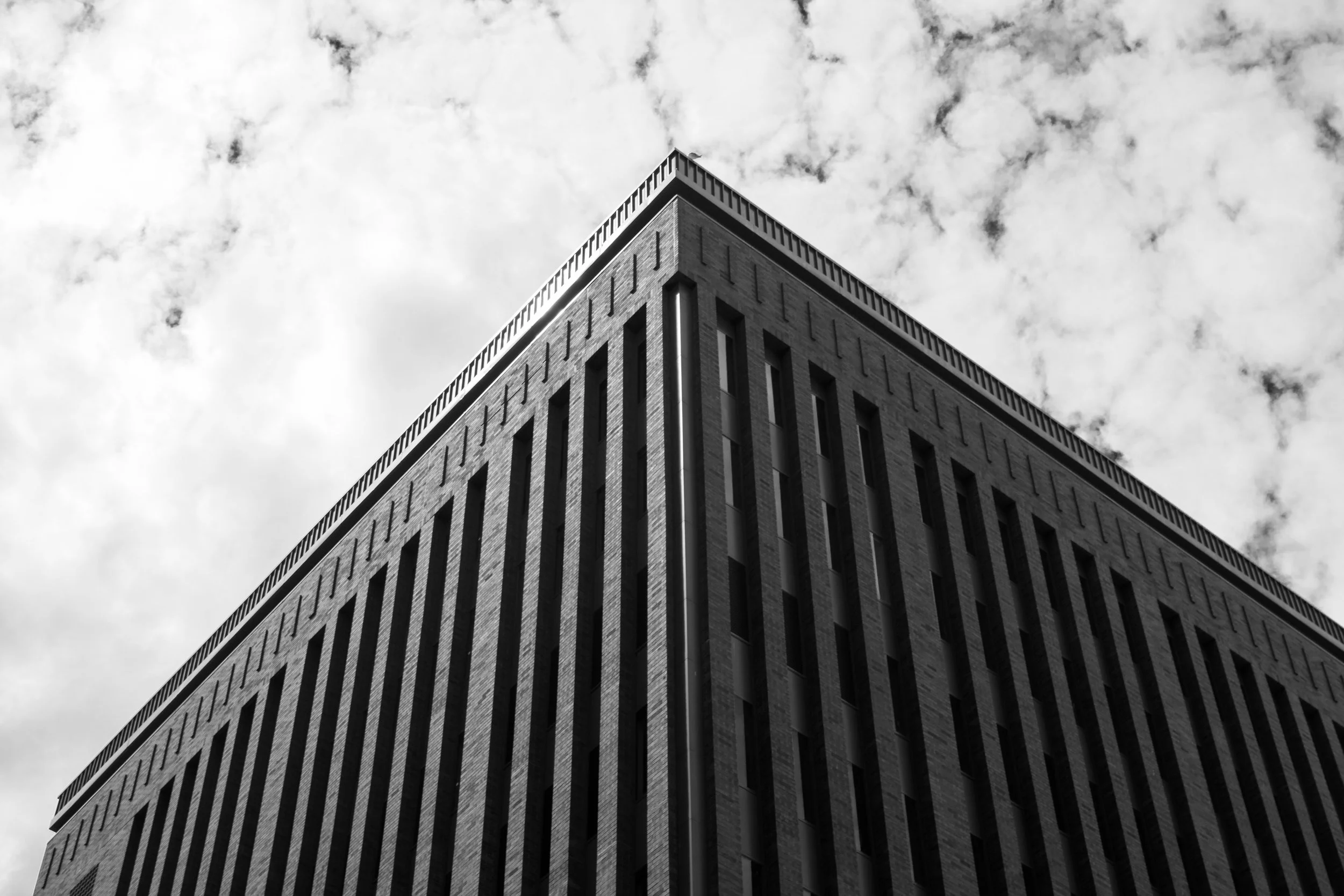 A tall, modern building with a flat roof and many rectangular windows, viewed from a low angle against a cloudy sky.