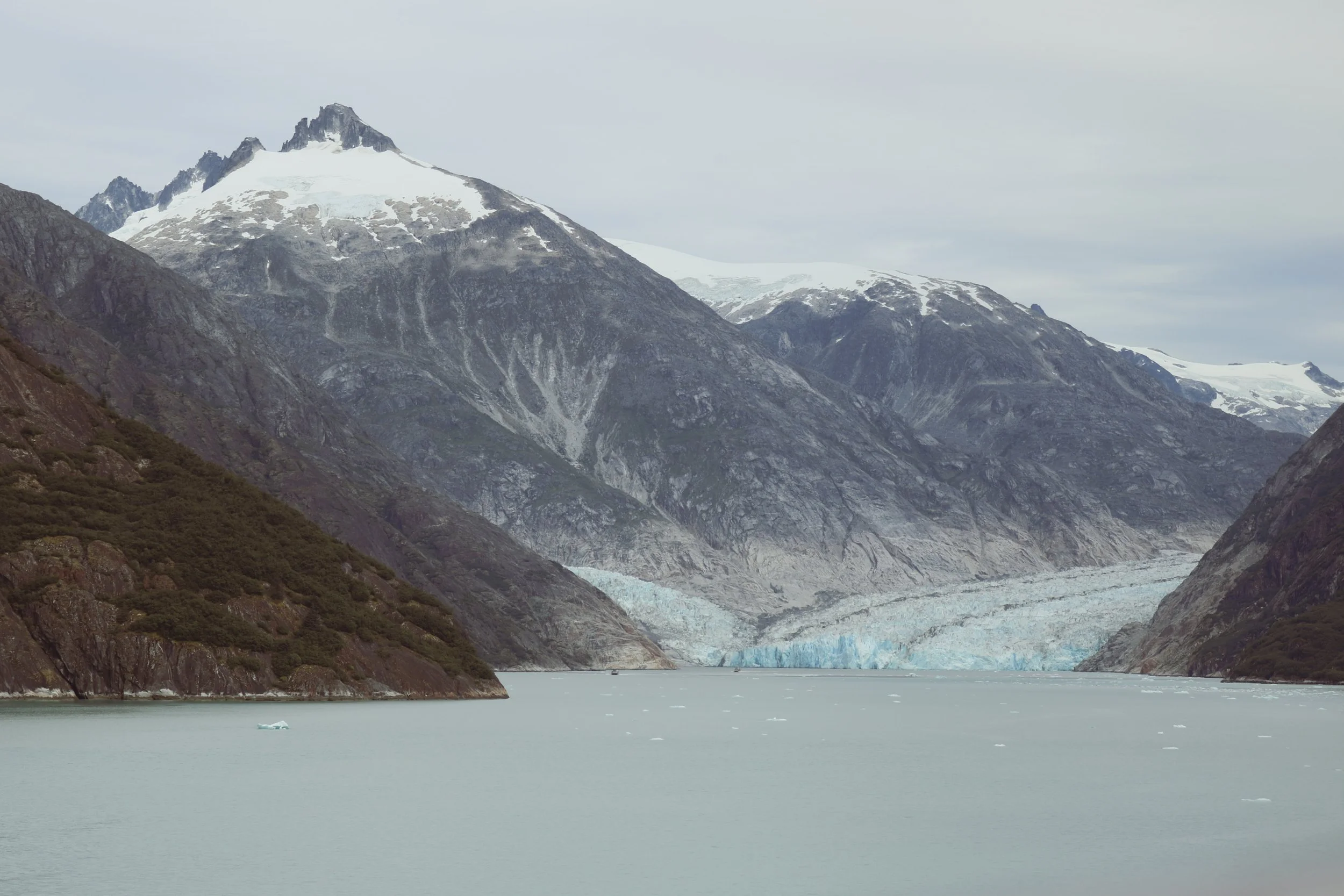 A glacier flowing from snow-capped mountains into a calm, icy body of water in a mountainous landscape.