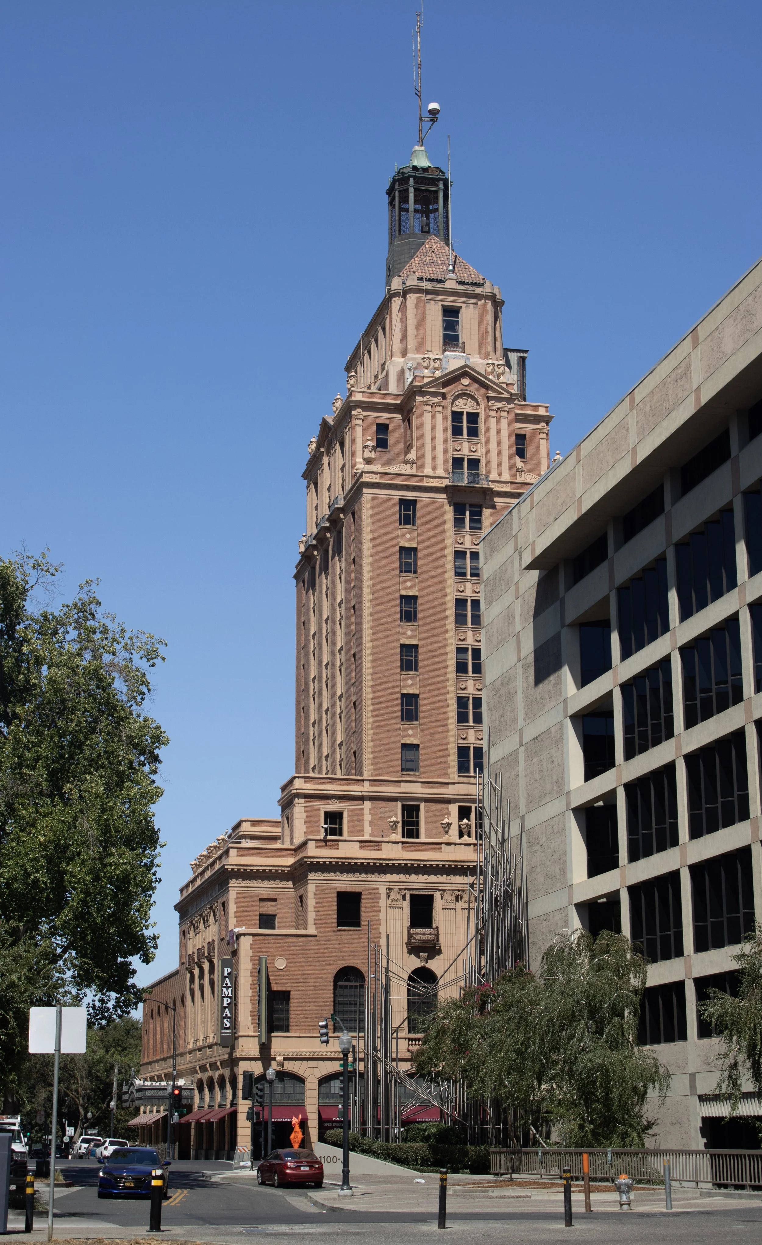 A tall, historic brick building with a clock tower and a flagpole, situated on a city street with cars, trees, and a modern office building nearby.