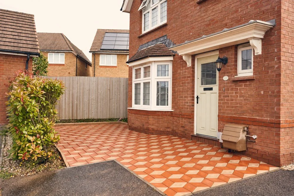 Front yard with patterned brick patio, a white front door with a window, a wall-mounted lantern, a window with shutters, a small window above, a beige pet house, and a wooden fence in the background.