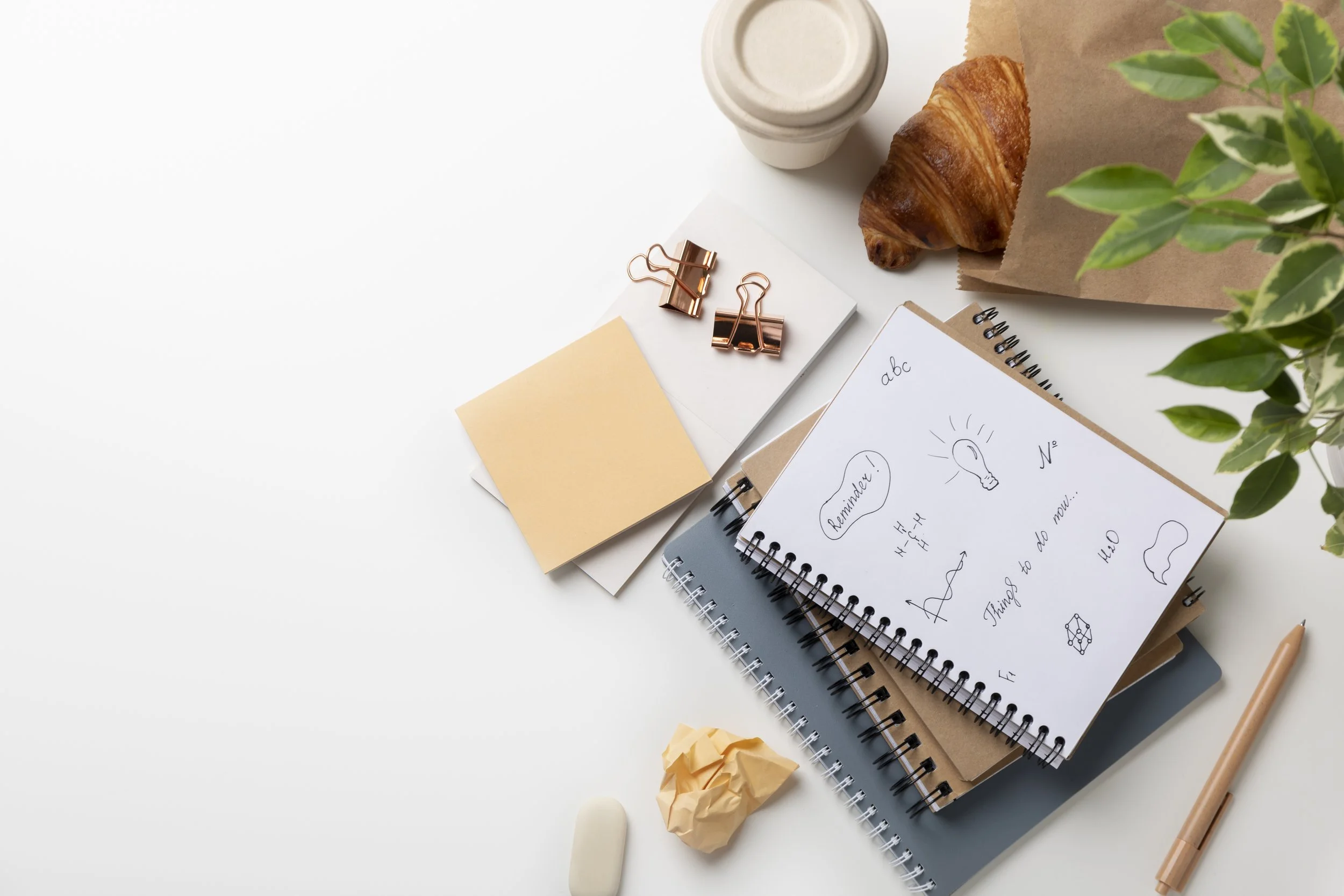 Flat lay of a workspace with notebooks, sticky notes, a crumpled piece of paper, a wooden pencil, a potted plant, and sunlight.