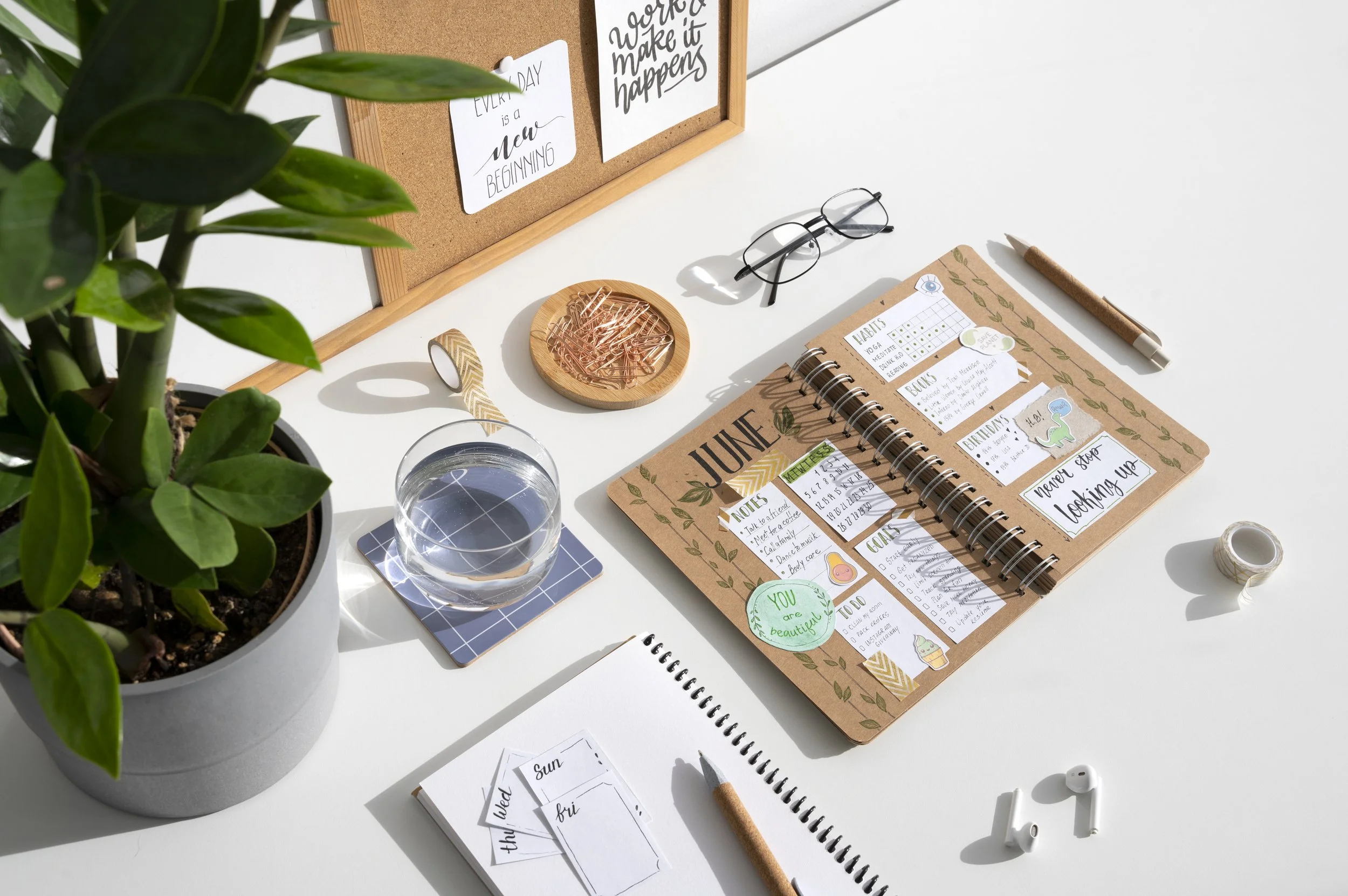 A desk workspace with a potted plant, glasses, a glass of water, a calendar, a notebook with handwritten notes, a corkboard with motivational quotes, and office supplies like paper clips, washi tape, and a pen.