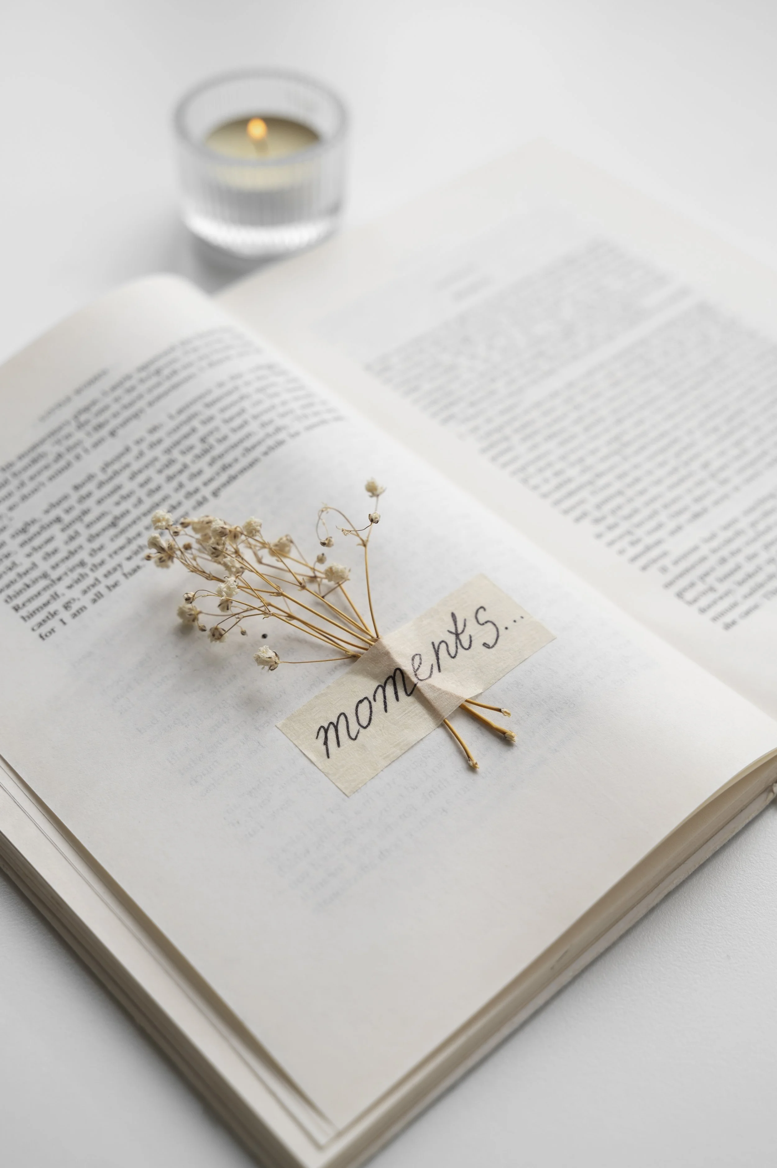 An open book with a small bouquet of dried white flowers labeled "moments" resting on its pages, and a lit candle in a glass holder placed behind the book on a white surface.