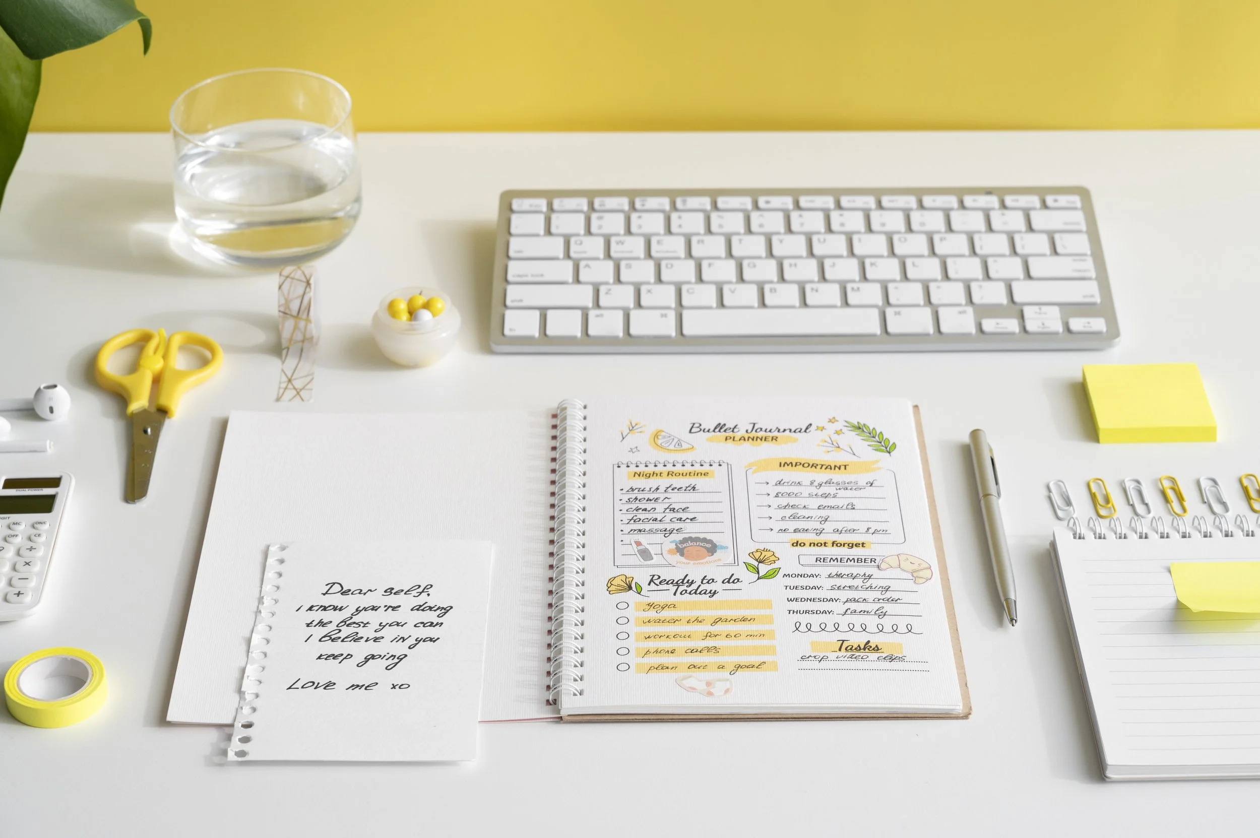 A white desk with a glass of water, white keyboard, yellow scissors, a small container with yellow and white candies, a notepad with handwritten notes and planner pages, a pair of white earphones, a yellow sticky note, and some yellow paper clips.