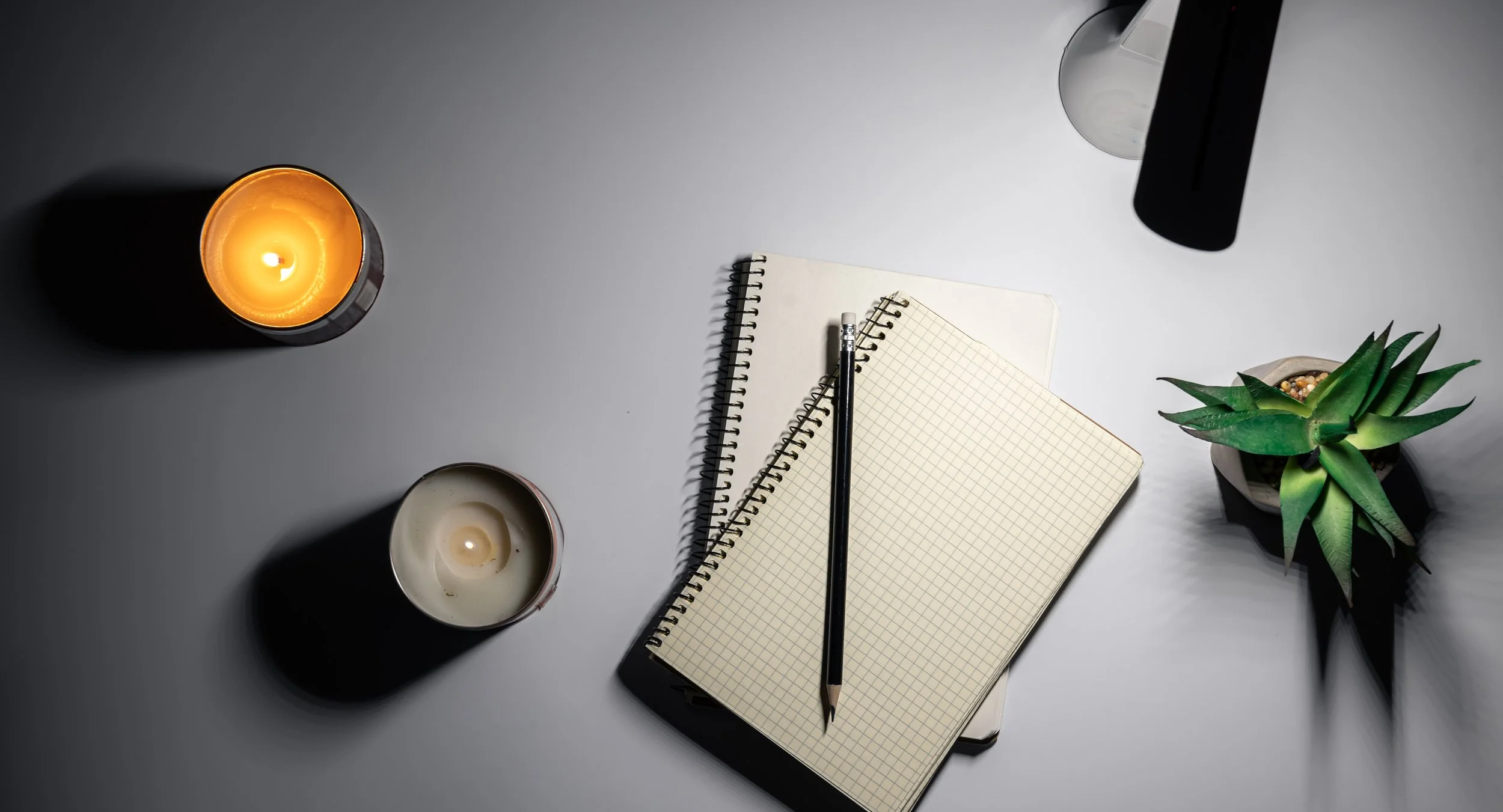 Overhead view of a desk with two candles, a spiral notepad, a grid paper notebook, an open black pen, a potted succulent, and a partially visible electronic device on a white surface.