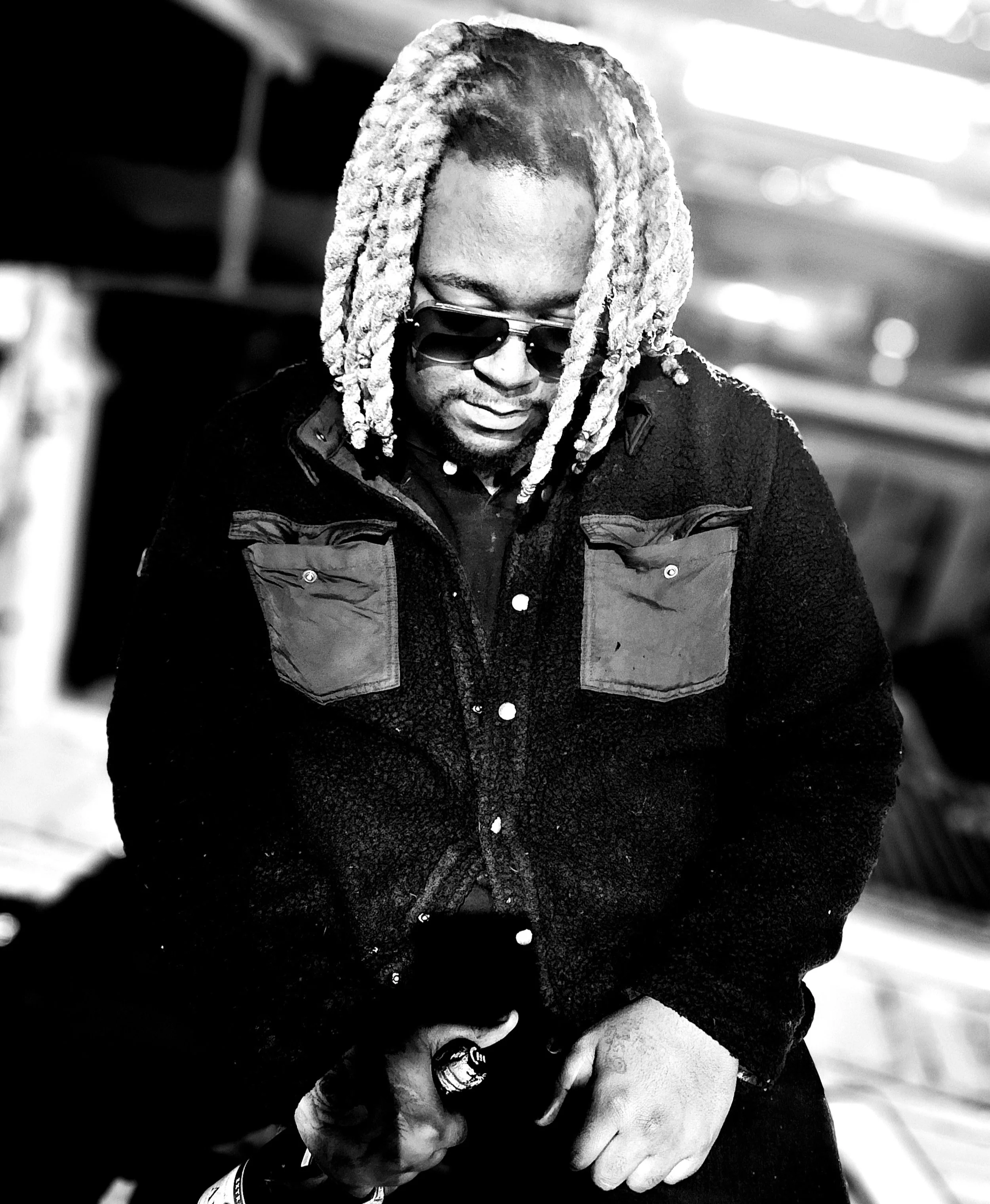 Black and white photo of a person with dreadlocks wearing sunglasses, comforting another person who is holding a microphone. The background appears to be indoors with blurred lighting.