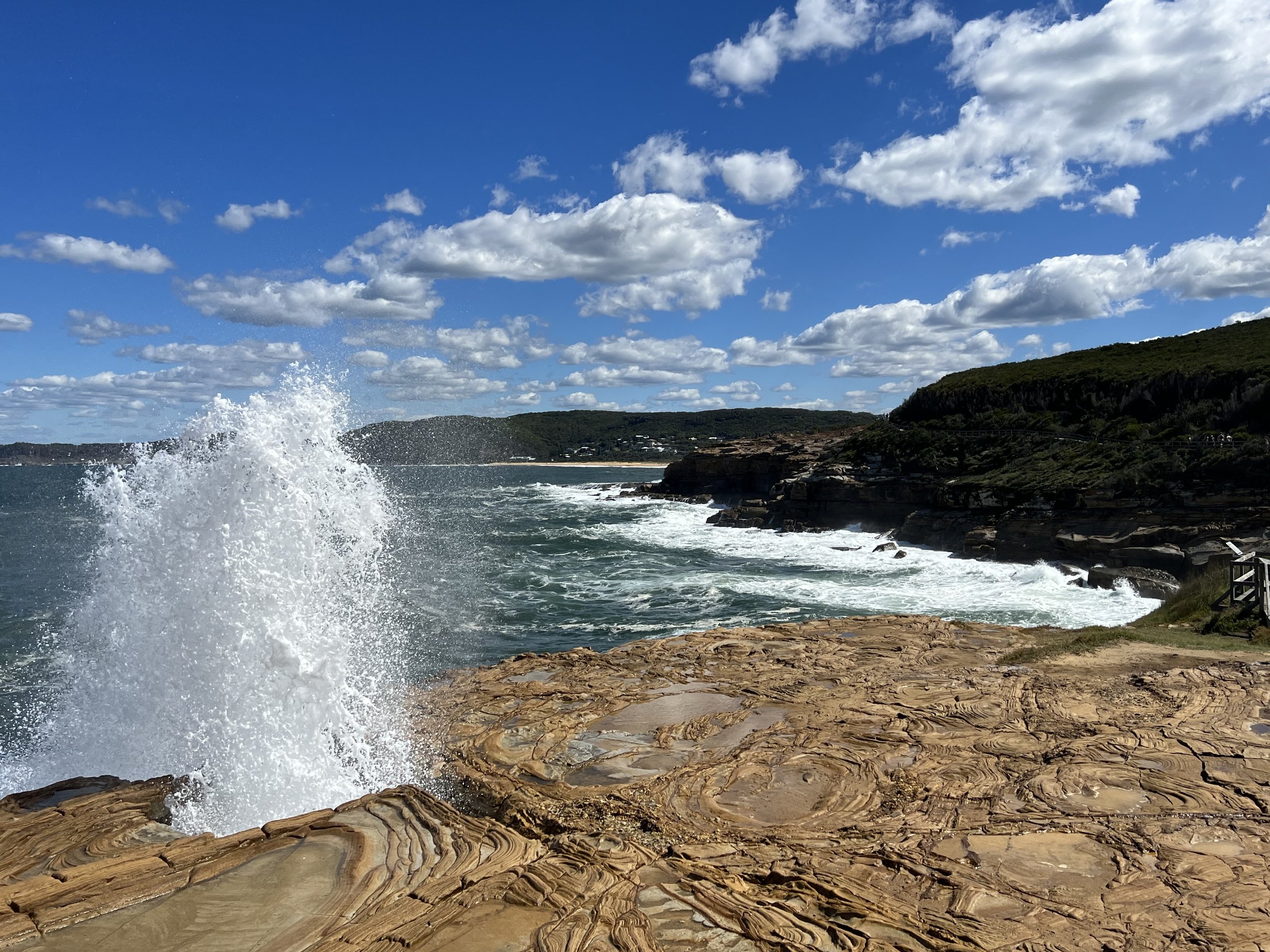 View of blowhole near Maitland Bay showing a rocky shoreline with ocean waves crashing, green hills in the background, and a partly cloudy sky.