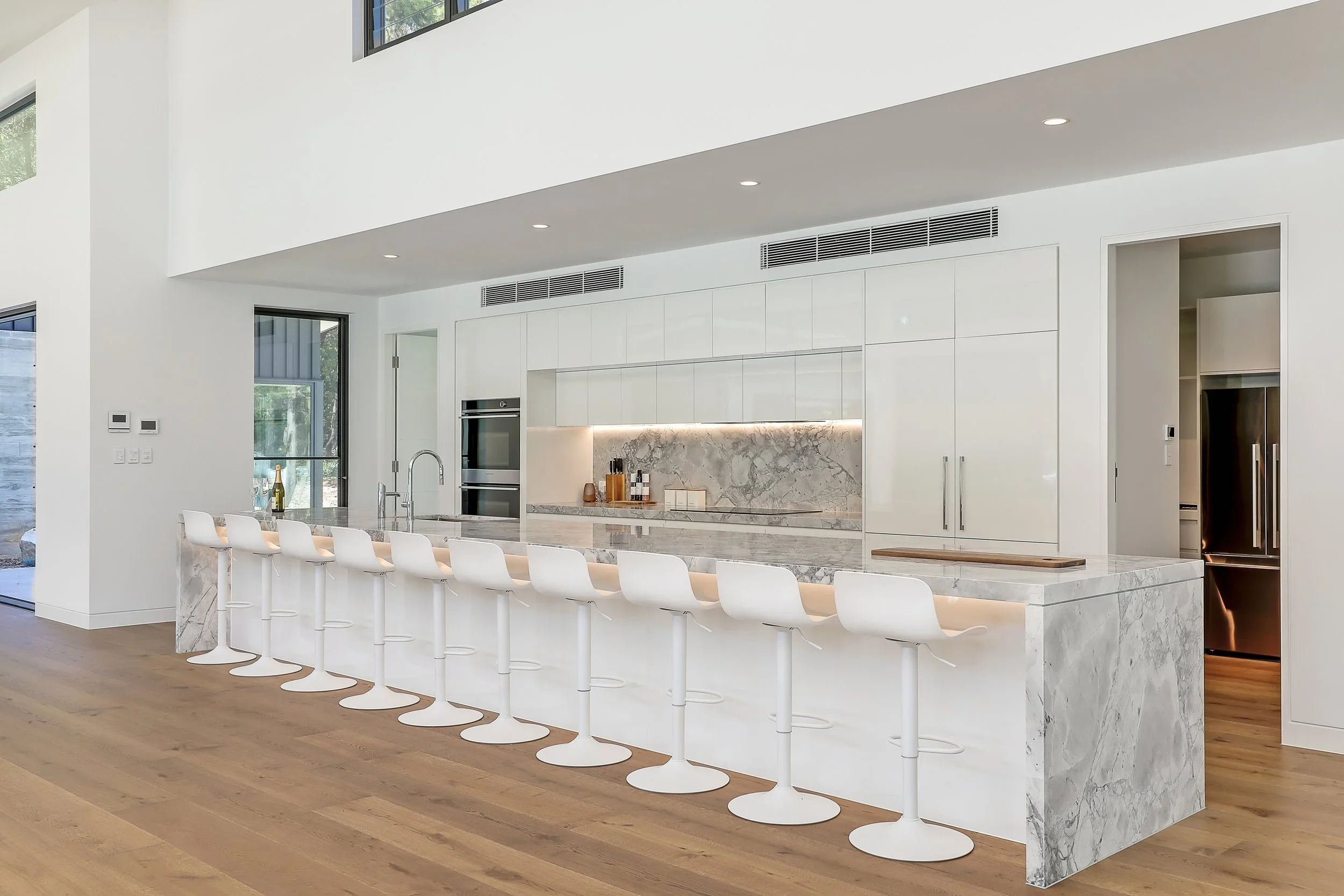 Launa's modern minimalist kitchen with white cabinetry, marble island, and white barstools, featuring a high ceiling and wood flooring.