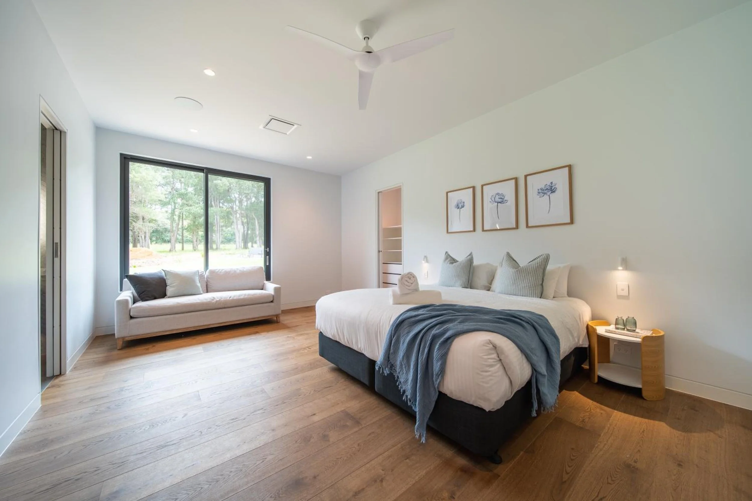 Another of Launa's modern bedrooms with a large window showing green outdoor scenery. The room features a white sofa, a bed with white bedding and blue accents, and framed floral artwork on the wall. 
