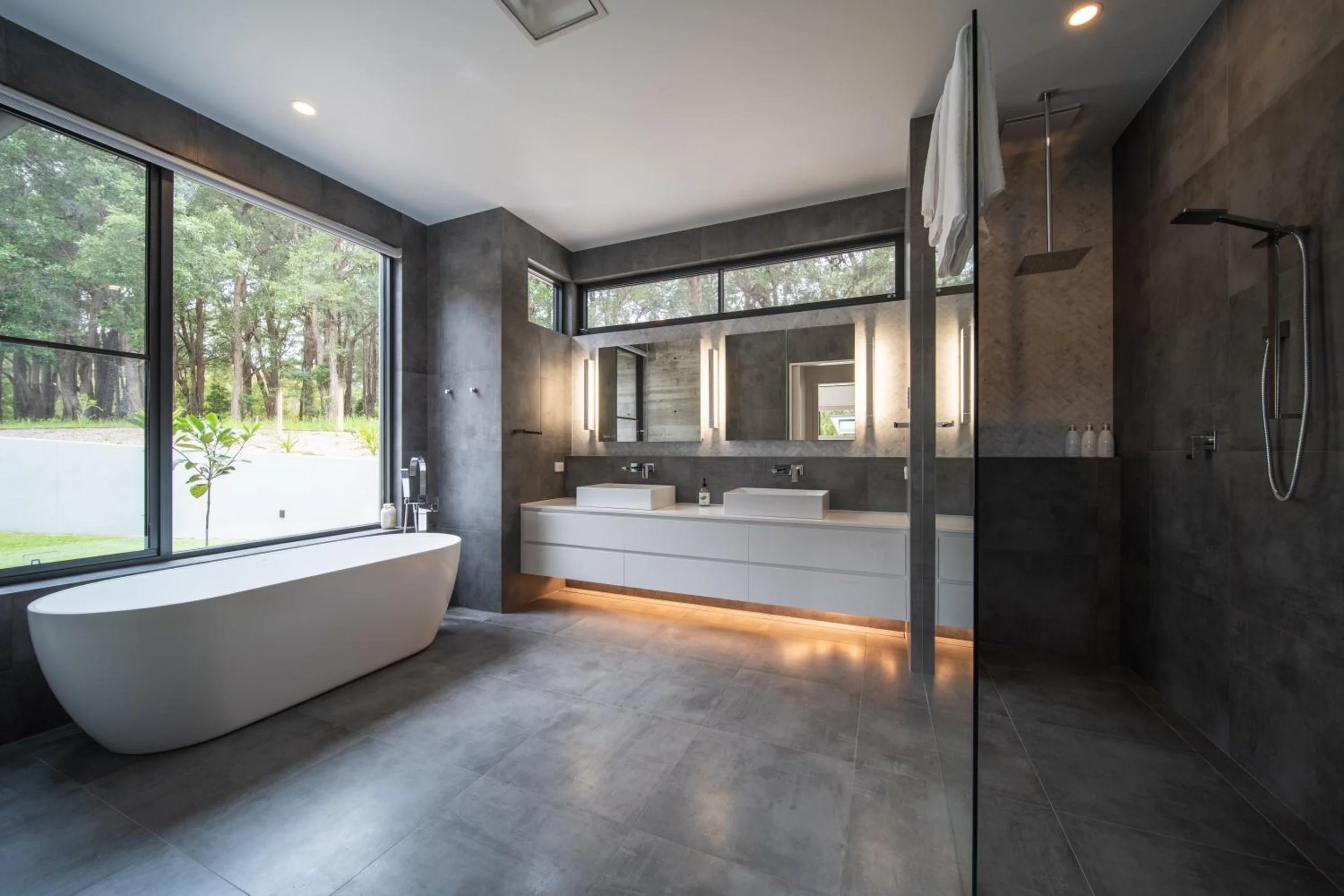 Another of Launa's modern bathrooms with large floor-to-ceiling windows showing trees outside, featuring a freestanding bathtub, double sink vanity, large mirror, and a rain shower head.