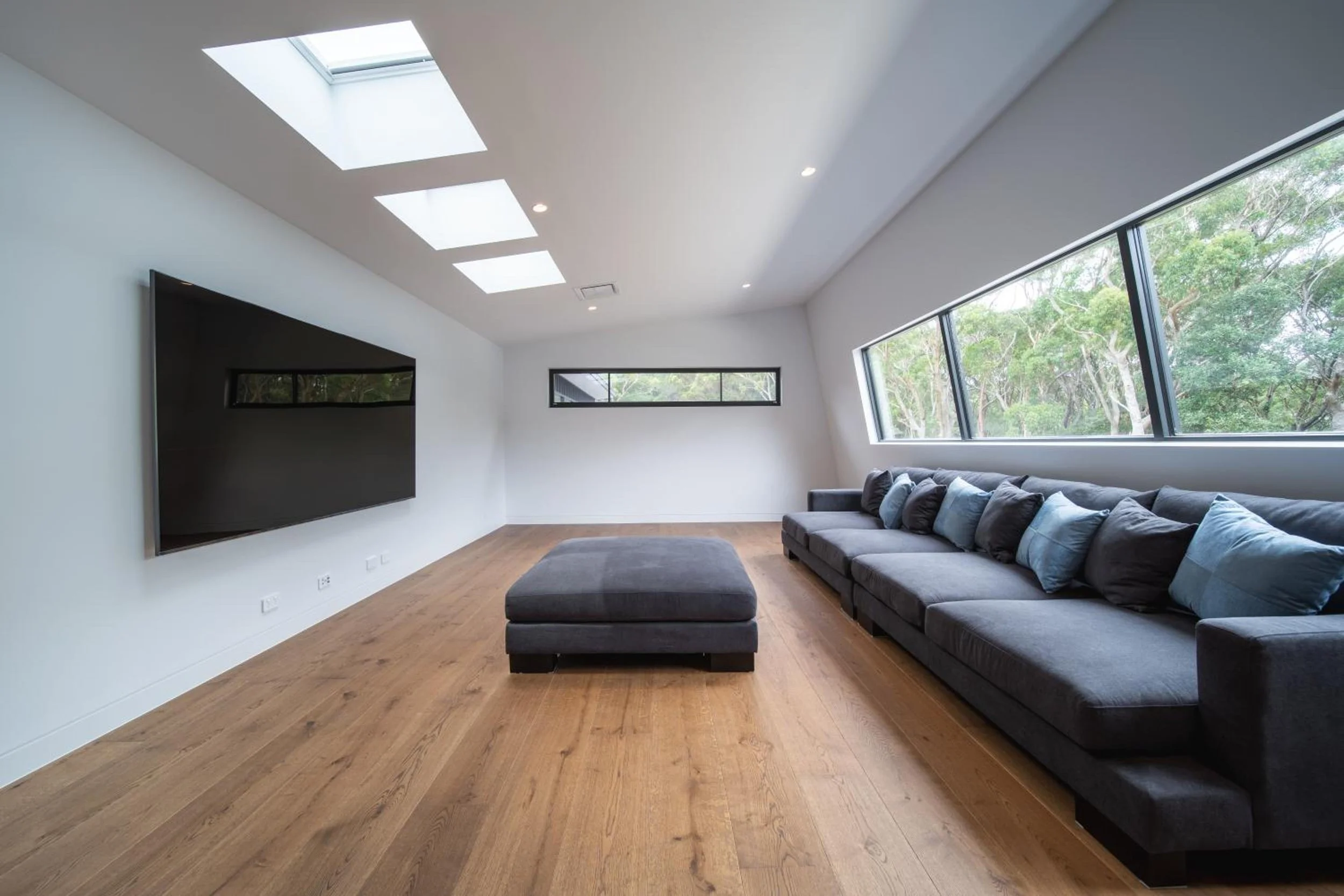 Another of Launa's modern living rooms with a large gray sectional sofa, black ottoman, flat screen TV on white wall, hardwood floor, large windows with green trees outside, and ceiling skylights.