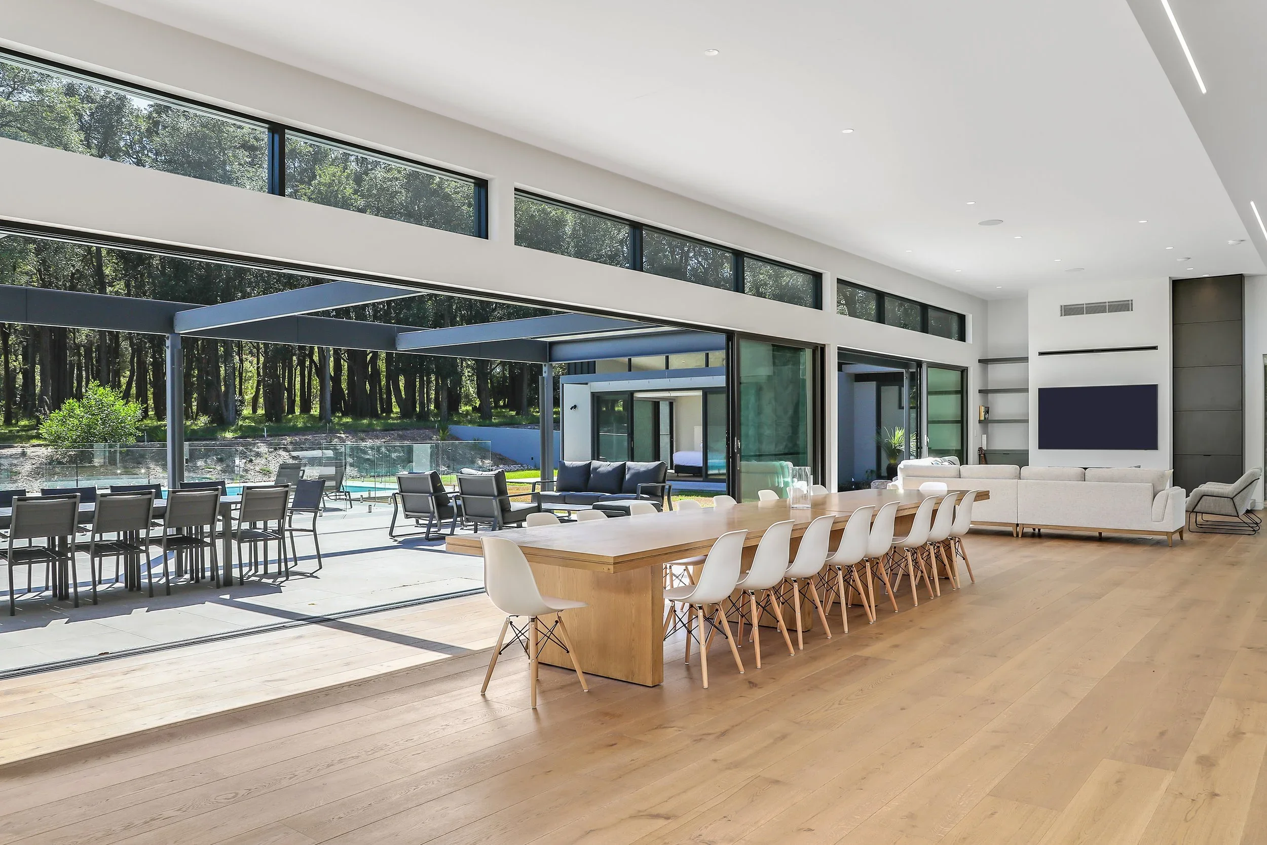 Launa's modern, spacious living and dining area with large floor-to-ceiling windows, white walls, and light wood flooring. There is an outdoor patio with chairs and a pool visible outside and the interior featuring a long wooden dining table.