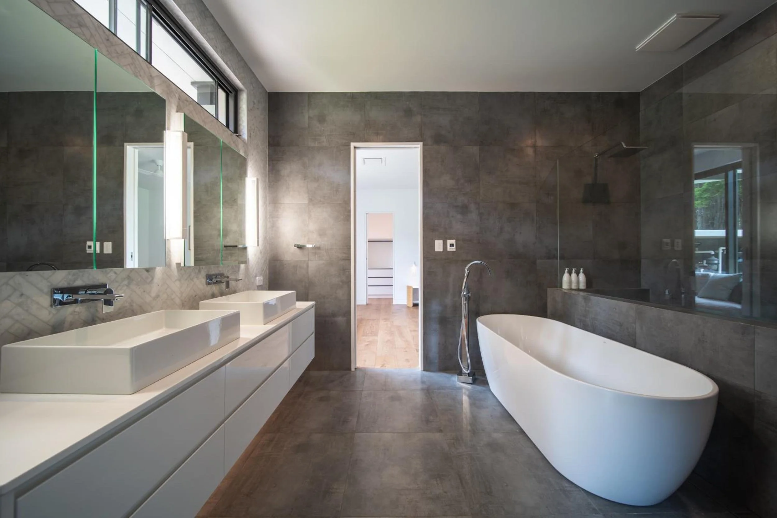 One of Launa's modern bathrooms with double white sinks, a large mirror, a freestanding bathtub, dark tiled walls, and a window with natural light.