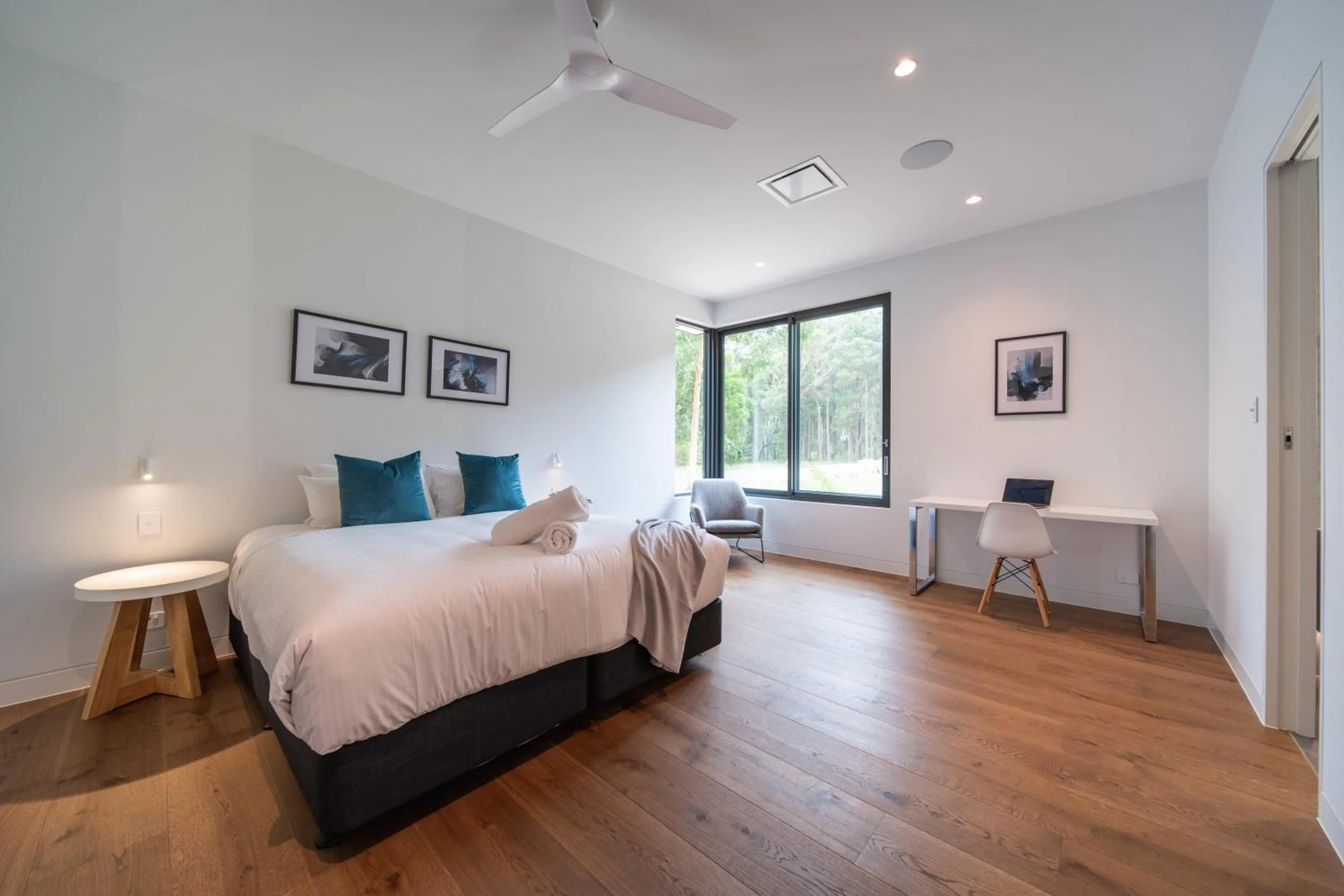 Another of Launa's bedrooms with large window, white walls, and wooden flooring. Contains a bed with a white comforter and blue pillows, a small wooden side table with a lamp, and a desk with a gray chair. Two framed art pictures hang on the wall.