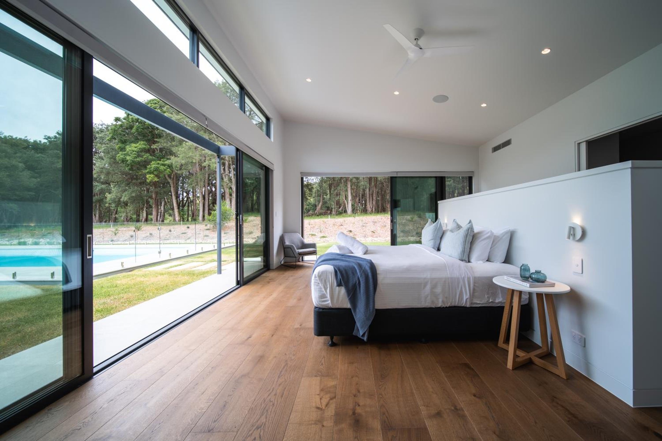 One of Launa's modern bedroom with large glass sliding doors opening to a green outdoor area, white bed with pillows and blanket, small side table with decorative items, light-coloured wooden floor, and ceiling fan.