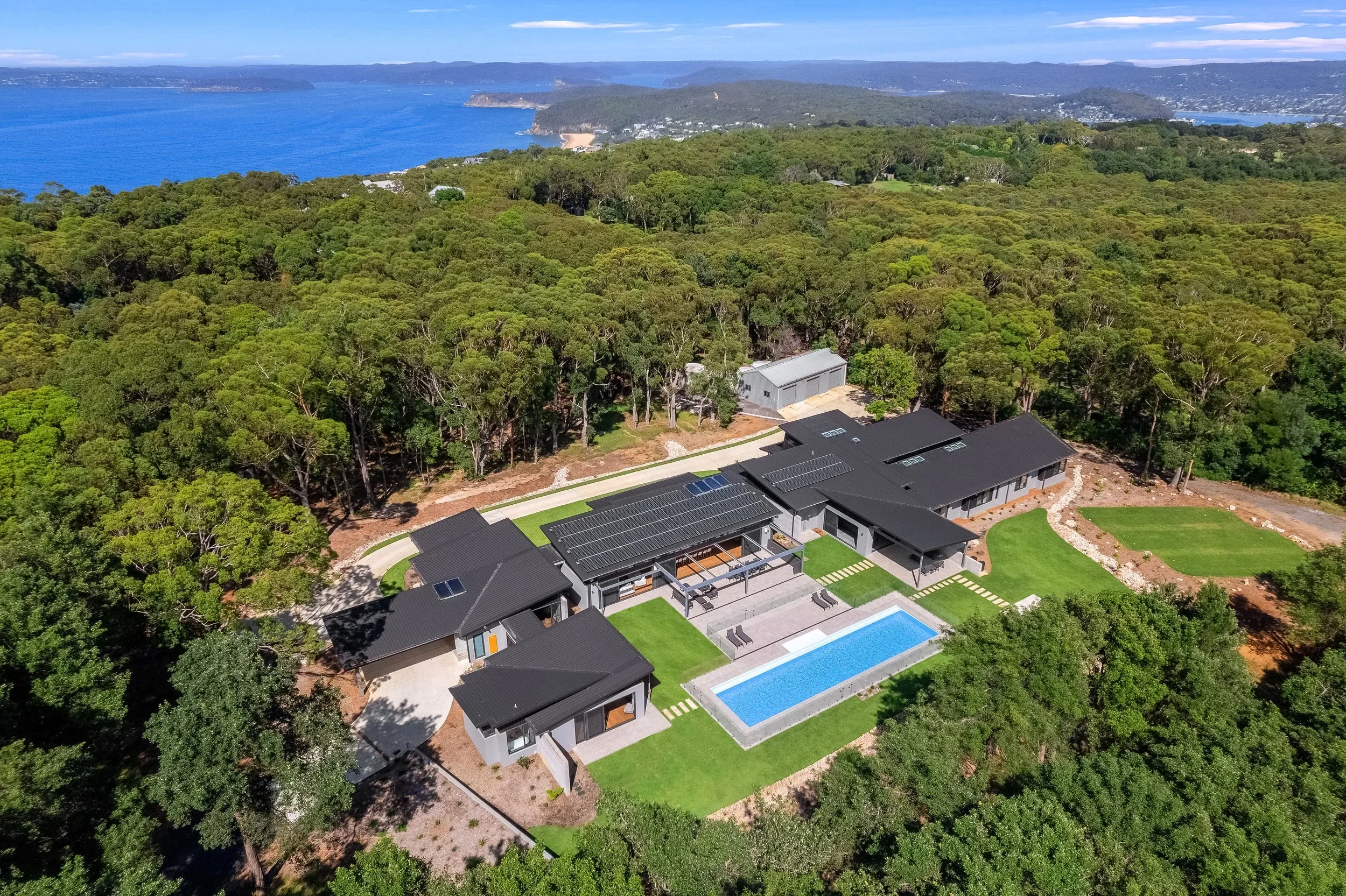 Aerial view of a Launa with solar panels and a swimming pool, surrounded by dense green trees, overlooking the ocean and distant hills.