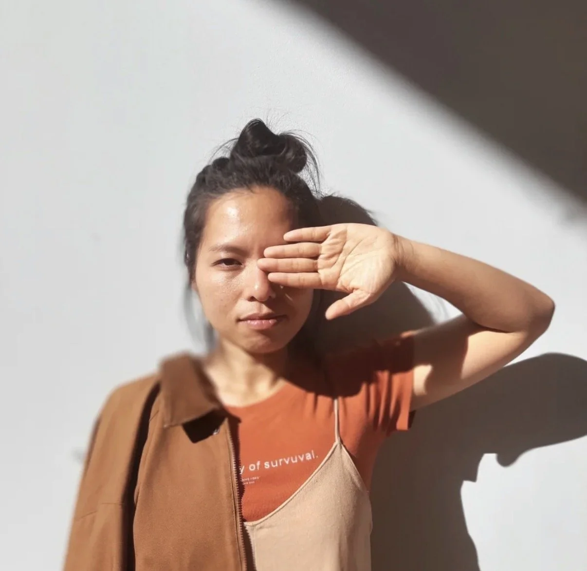 Khanh Doan, chef, with dark hair in a messy bun, standing against a white wall, shielding her right eye with her hand, wearing an orange t-shirt and a brown jacket.