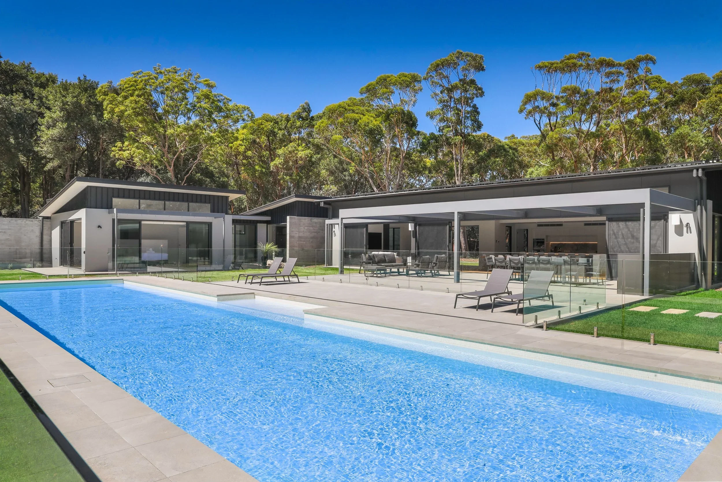 Launa with large glass windows and sliding doors, outdoor patio with furniture, a swimming pool, green lawn, surrounded by tall trees and a clear blue sky.
