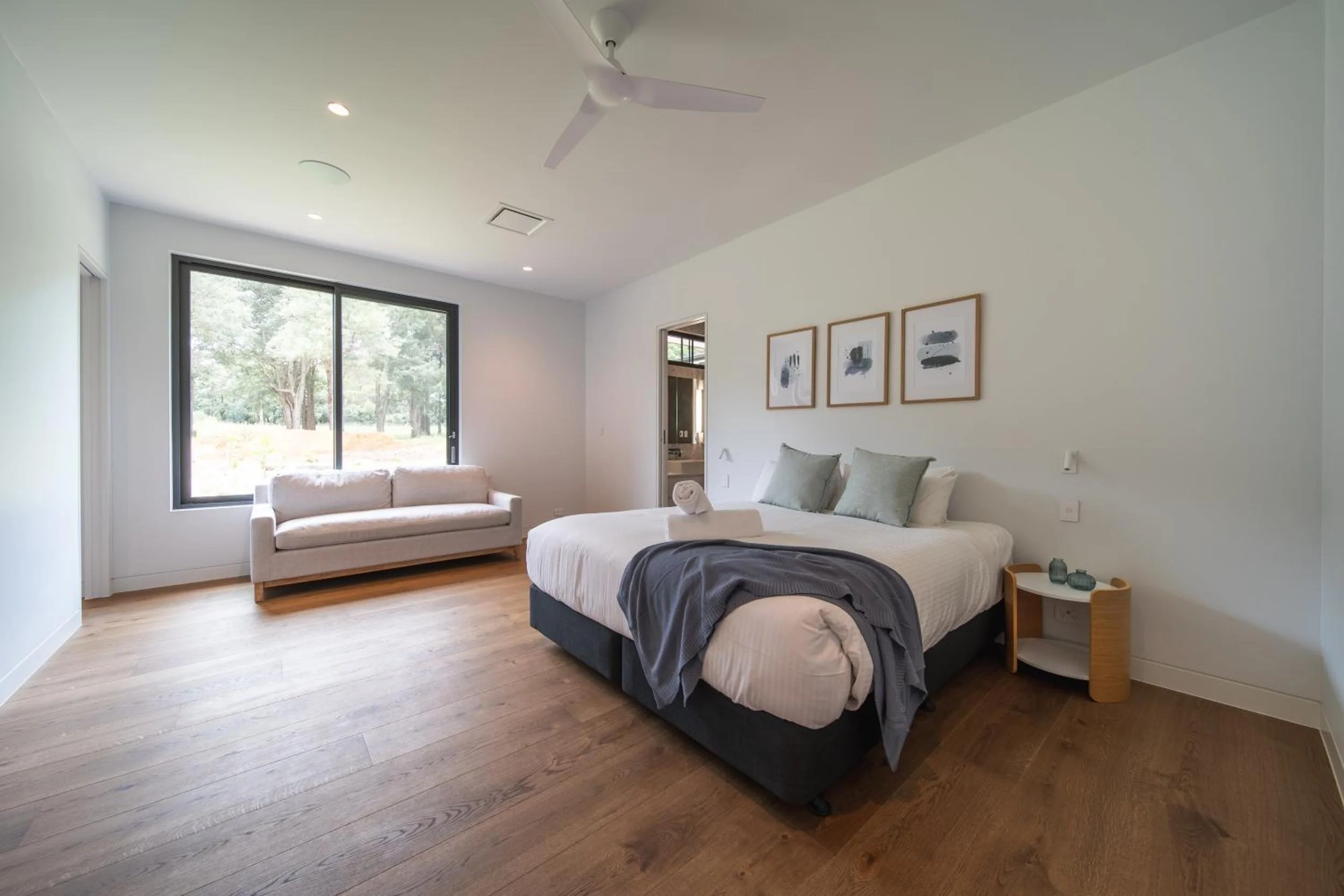 A bedroom with a large window showing green trees outside, a bed with gray and white bedding, a small round side table with decorative items, a beige loveseat, framed artwork on the wall, and wooden flooring.