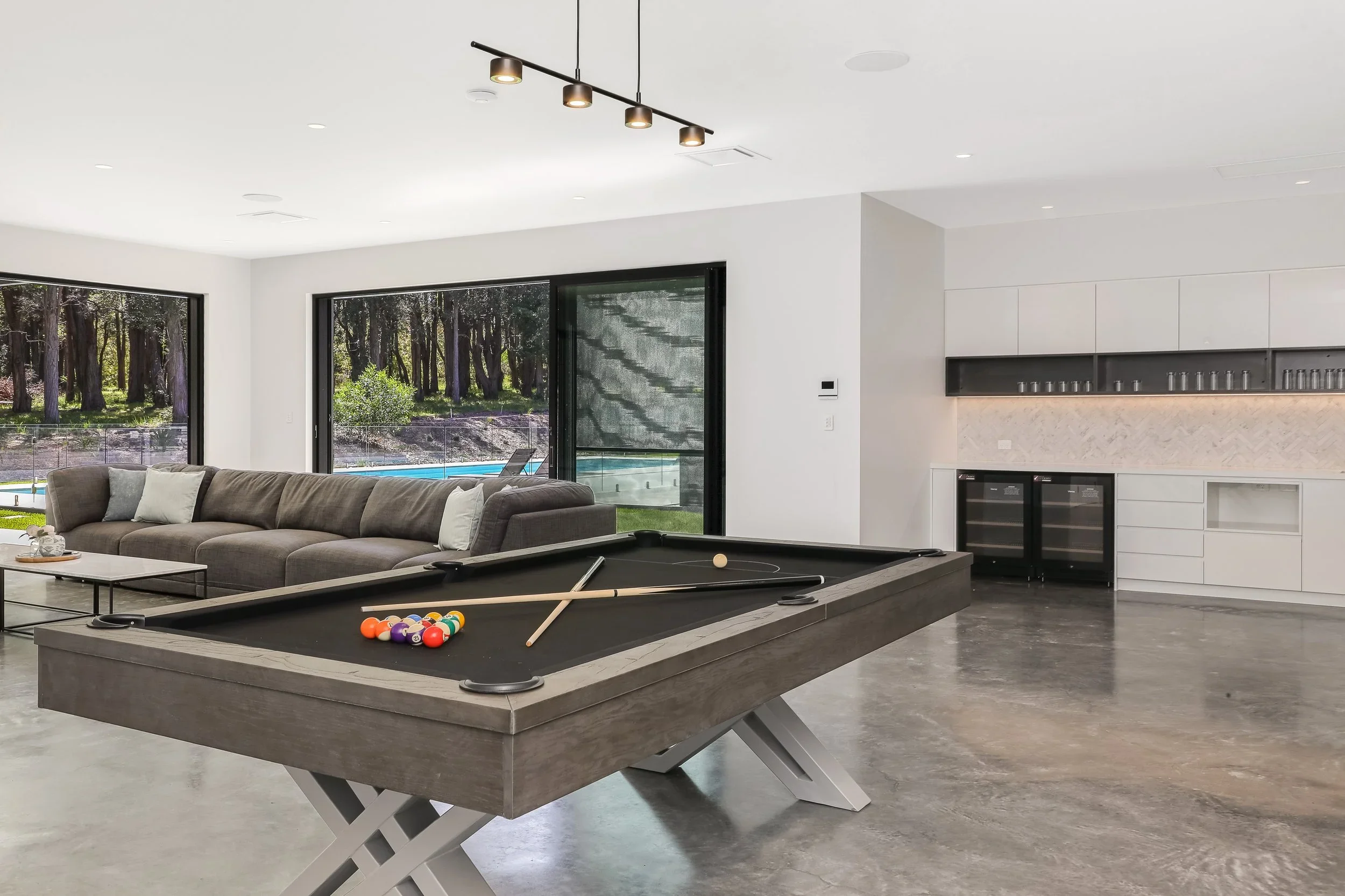 Another of Launa's modern living room with pool table, gray sofa, large windows showing outdoor trees and pool, white walls, and minimalist decor.