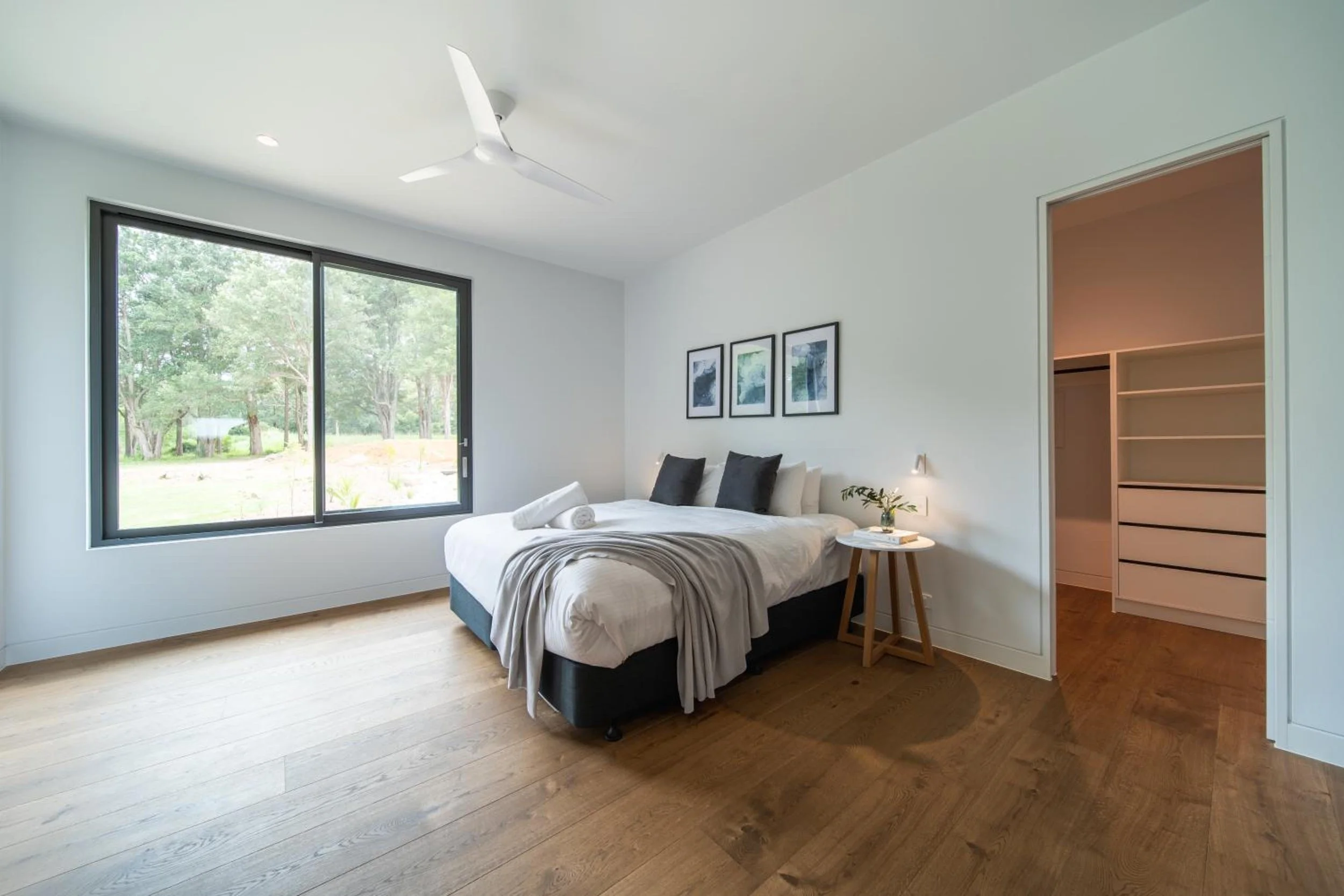 Another of Launa's bedroom with a large window showing trees outside, a bed with white bedding and black pillows, a side table with a small plant, artwork on the wall, and an open closet with shelves.