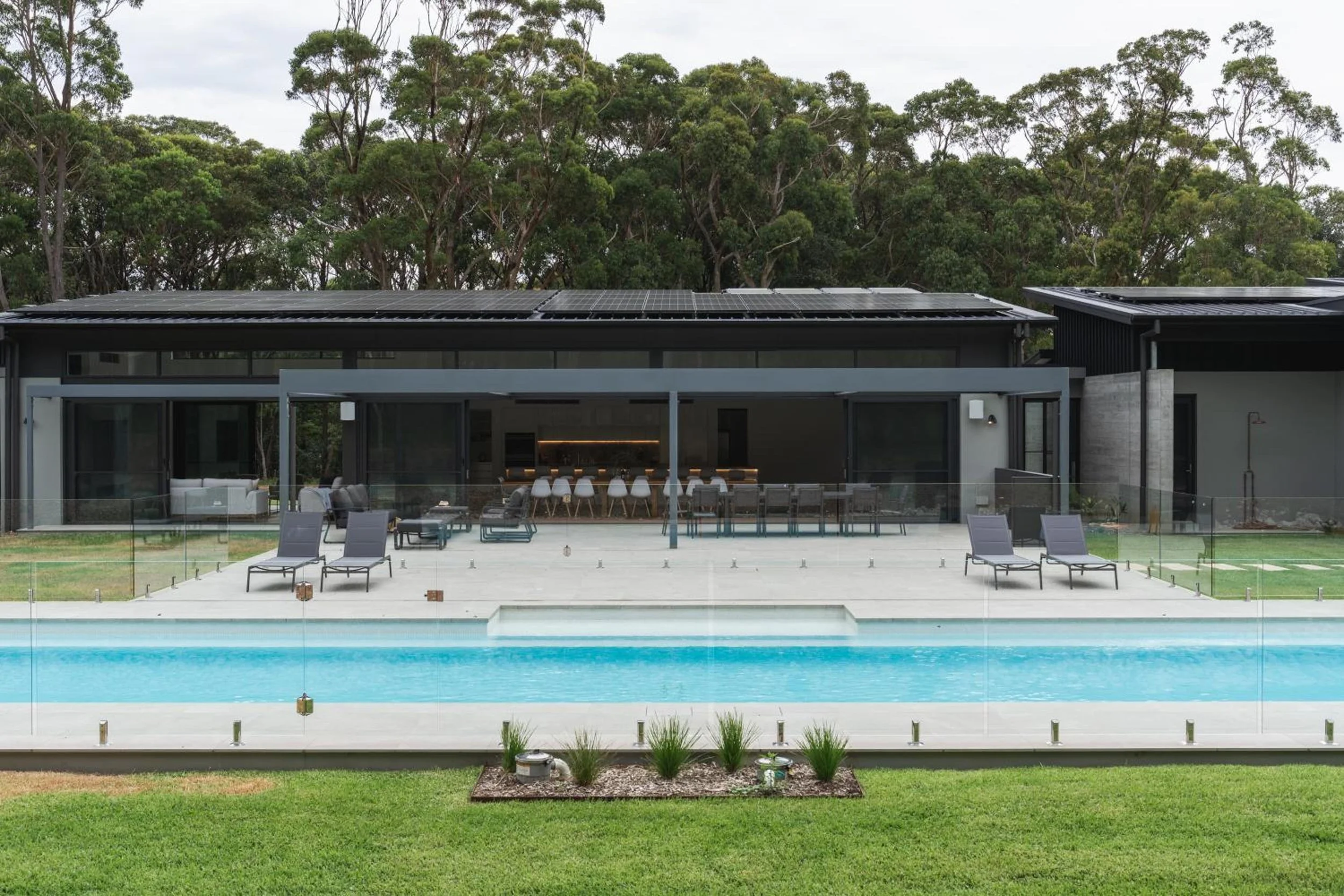 View of Launa house with solar panels on roof, large glass windows, outdoor patio with lounge chairs, swimming pool with blue water, and surrounded by green lawn and trees.