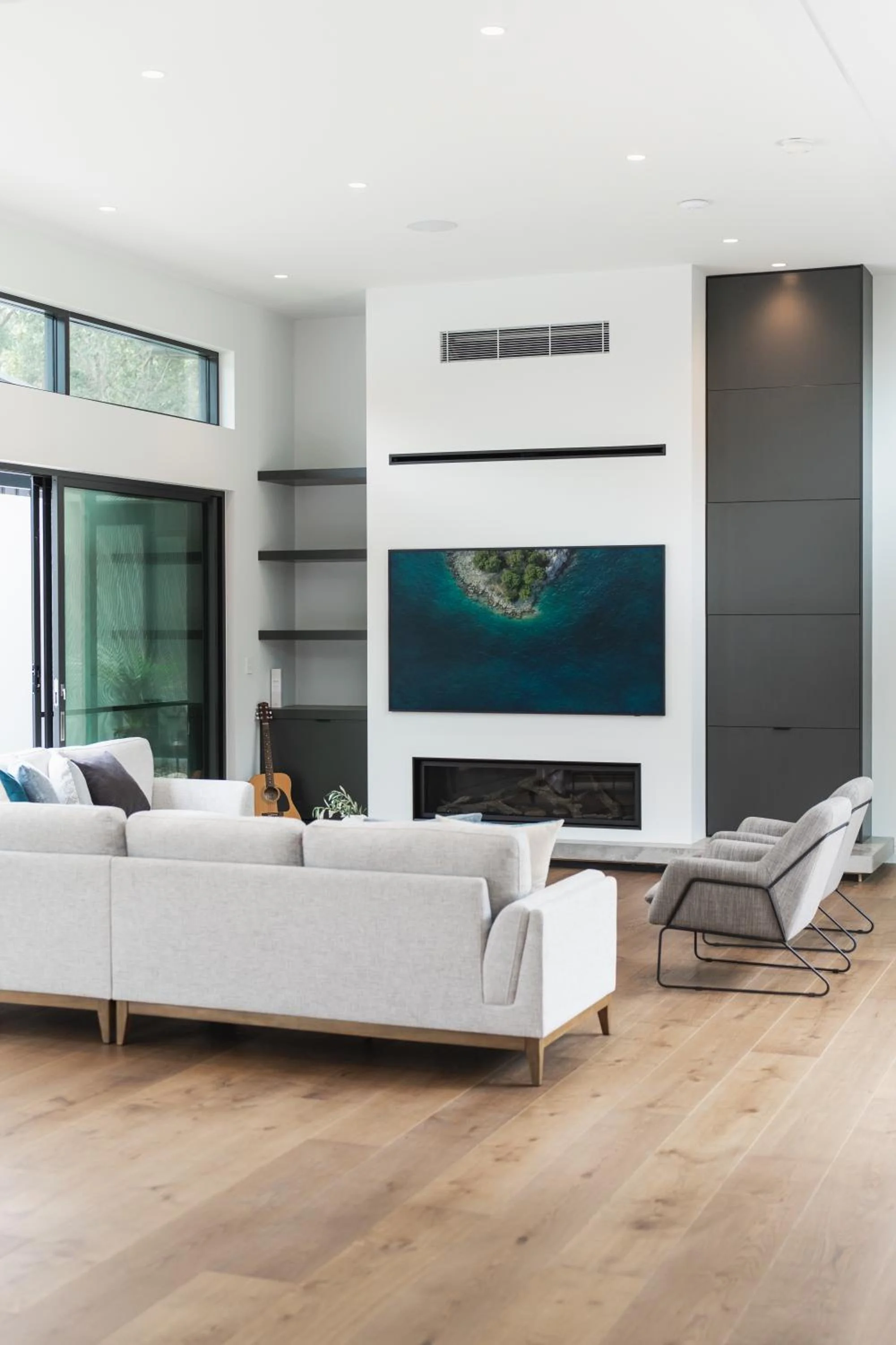 Launa's modern living room with white sofa, a gray armchair, a fireplace, wall-mounted TV showing an aerial view of a small island with greenery surrounded by water, large windows, and wooden flooring.