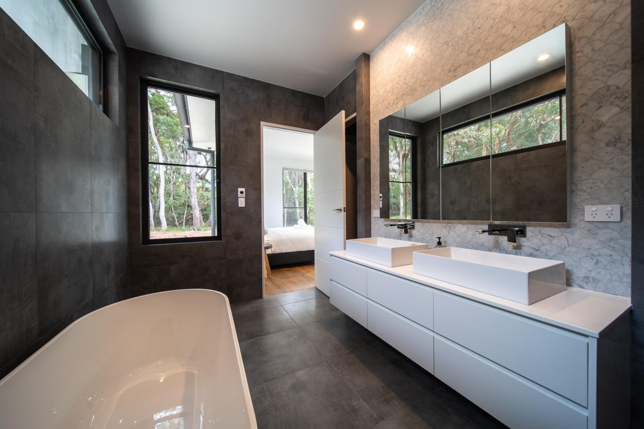 Another of Launa's modern bathrooms with double sinks, large mirror, tiled walls, and large windows showing trees outside.