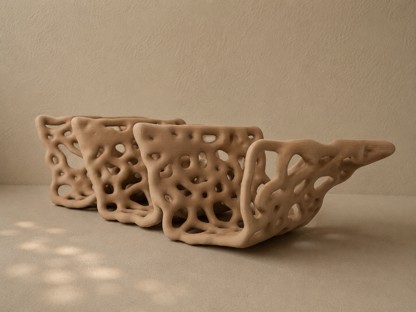 Three beige, abstract, openwork ceramic bowls with irregular holes, placed on a beige surface against a beige wall.