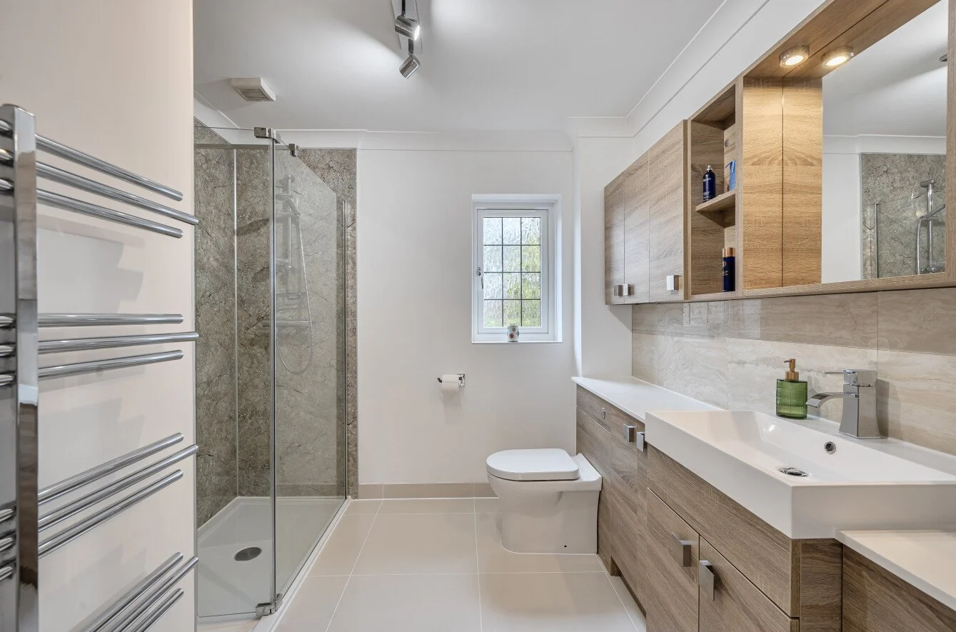 Modern bathroom with walk-in shower, wooden cabinetry, white sink, toilet, and small window.