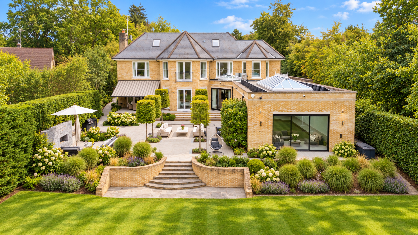 Spacious backyard of a large two-story brick house with a patio, outdoor seating, lush garden, manicured lawn, and surrounding tall trees under a partly cloudy sky.