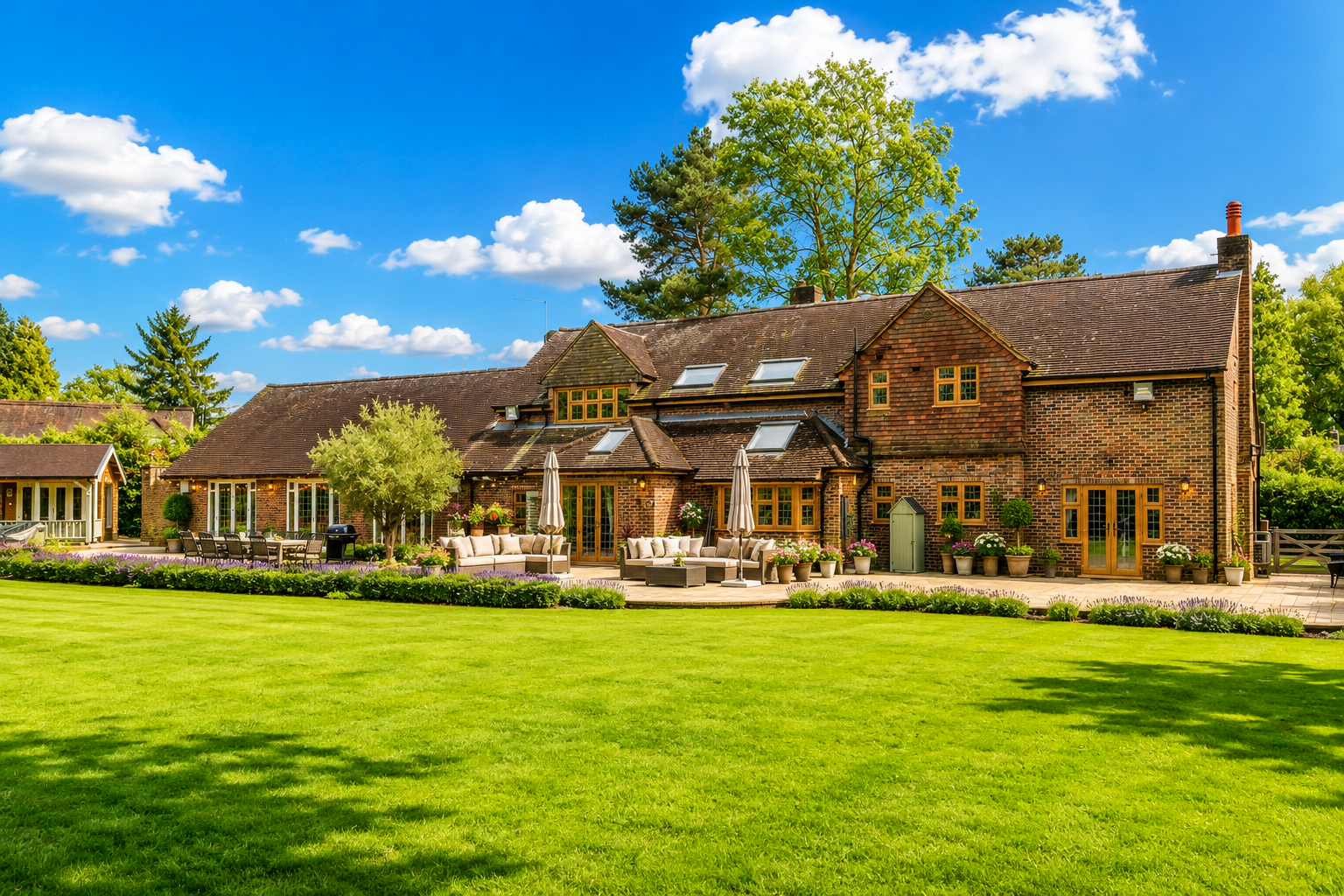 Large brick house with multiple windows and skylights, surrounded by a well-maintained lawn and garden with patio furniture, potted plants, and umbrellas, under a bright blue sky with scattered clouds.