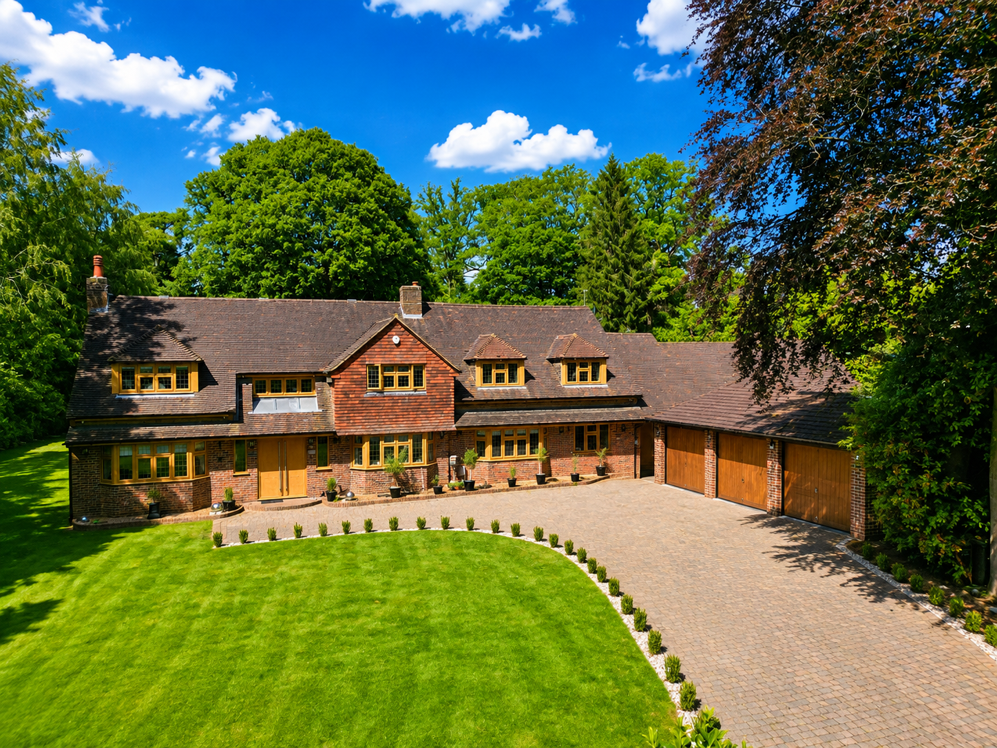 A large, two-story brick house with a brown tiled roof, multiple dormer windows, and a spacious driveway leading to a three-car garage. The house is surrounded by a well-maintained lawn and tall green trees under a partly cloudy blue sky.