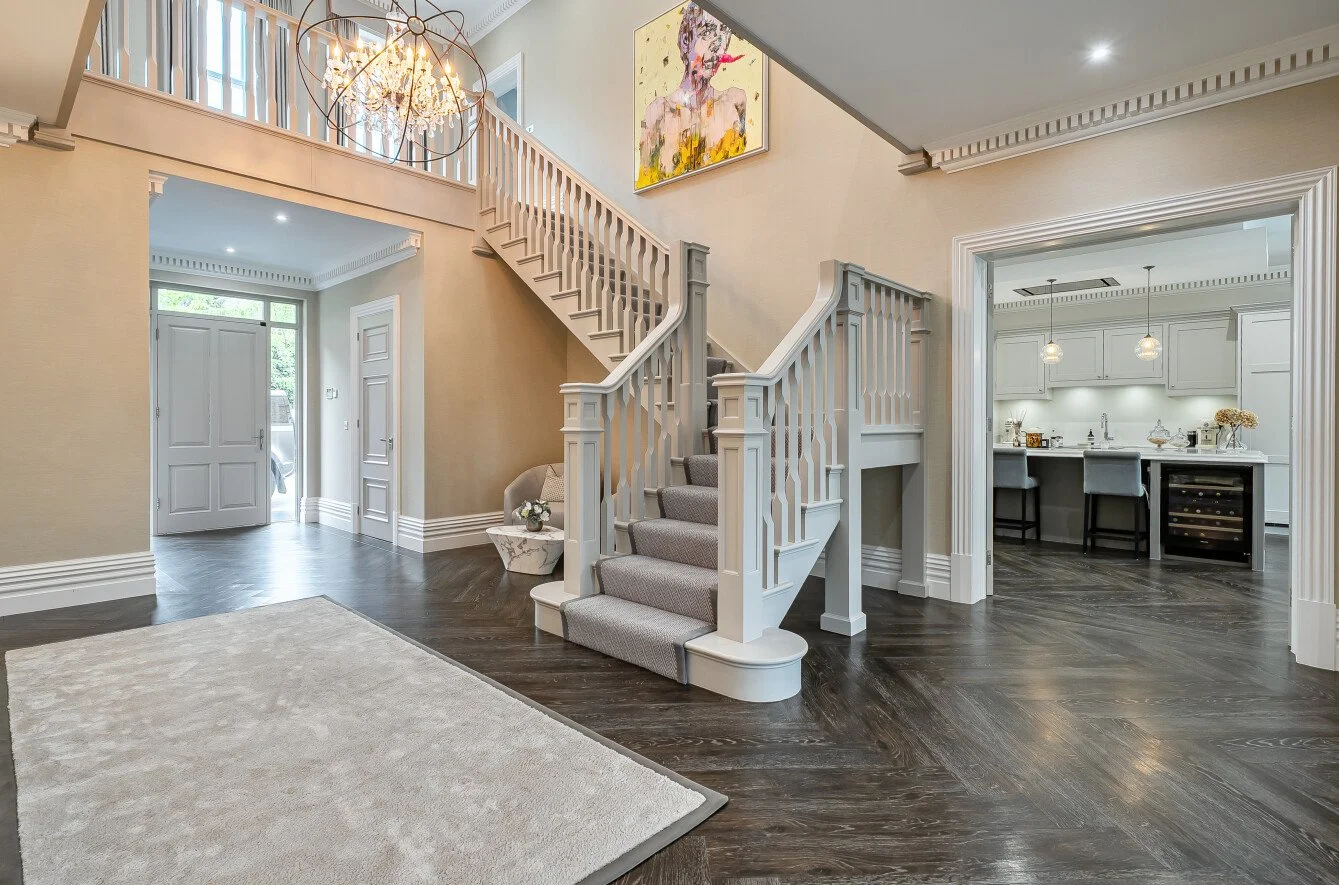 Entryway with staircase leading to upper level, chandelier hanging from ceiling, beige walls, dark hardwood flooring, and a hallway opening to the kitchen. A piece of modern art is displayed on the wall above the staircase.