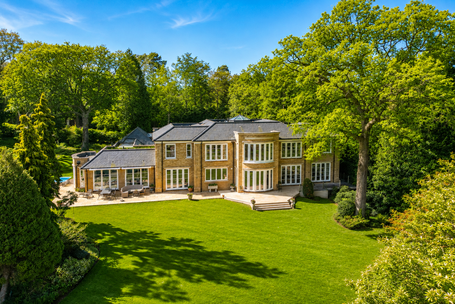 Large mansion with multiple levels and large windows, surrounded by lush green trees and a well-maintained lawn, under a clear blue sky.