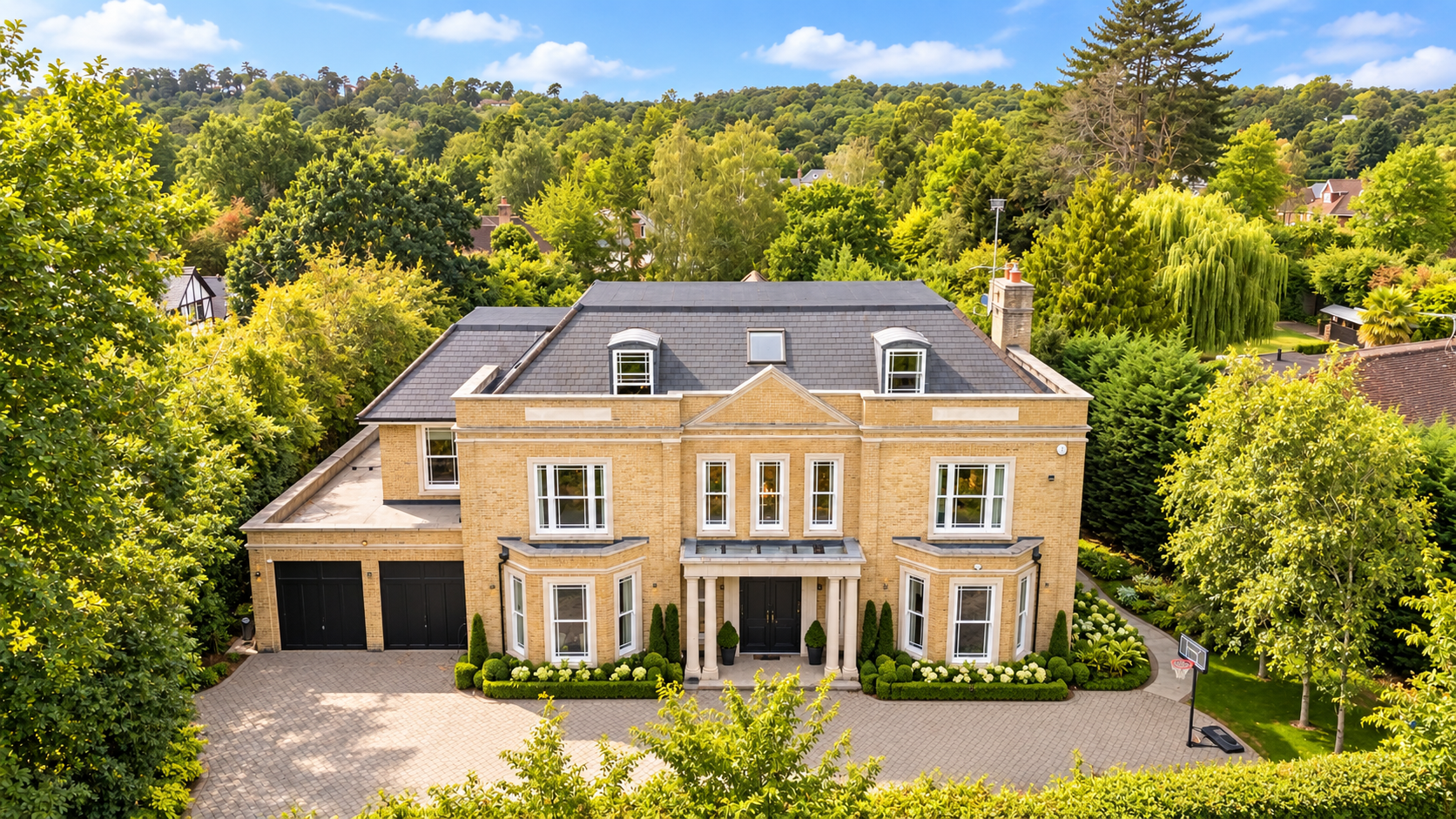 Large, two-story house with a beige brick exterior, black front door, and symmetrical windows. Surrounded by greenery, trees, and landscaped gardens. A paved driveway leads to a double garage, with a basketball hoop nearby. Hills and more trees in th