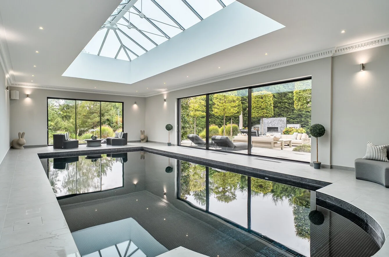 Indoor swimming pool in a modern home with large glass sliding doors opening to a patio with outdoor furniture and lush greenery.
