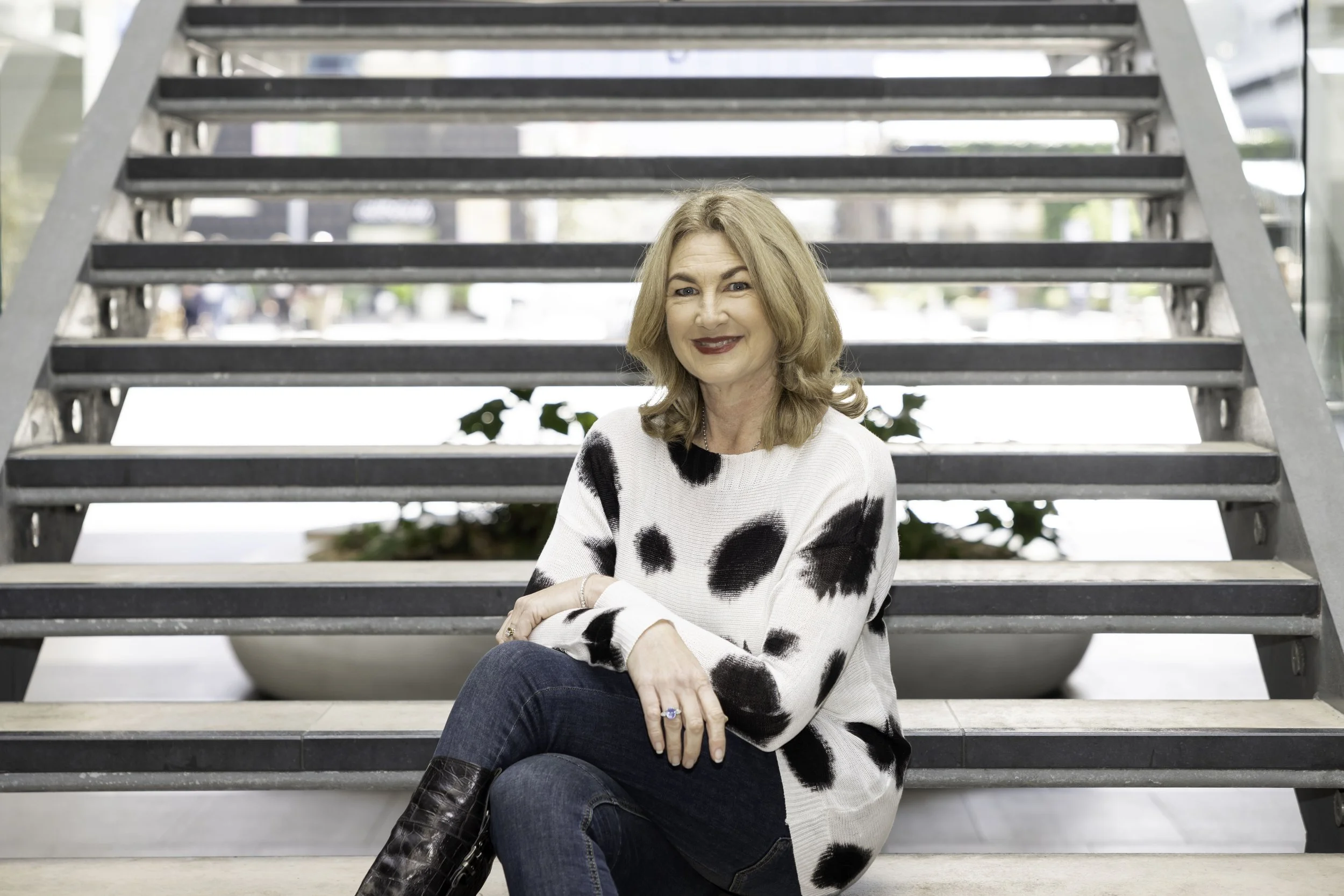A smiling woman with shoulder-length blond hair, wearing a white sweater with black spots and dark jeans, sitting on stairs in front of a modern building with large glass windows.