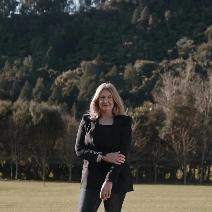 A woman with shoulder-length blonde hair standing outdoors in front of trees and mountains, smiling, wearing a black blazer and black pants.