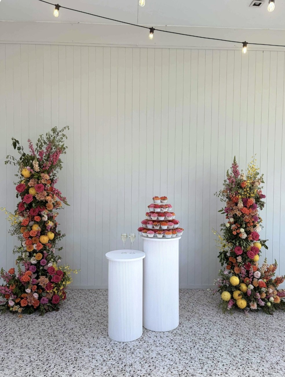 Decorative display with two tall floral arrangements, a tiered tower of cupcakes, and two glasses of champagne on white cylindrical pedestals, set against a white wall with string lights overhead.