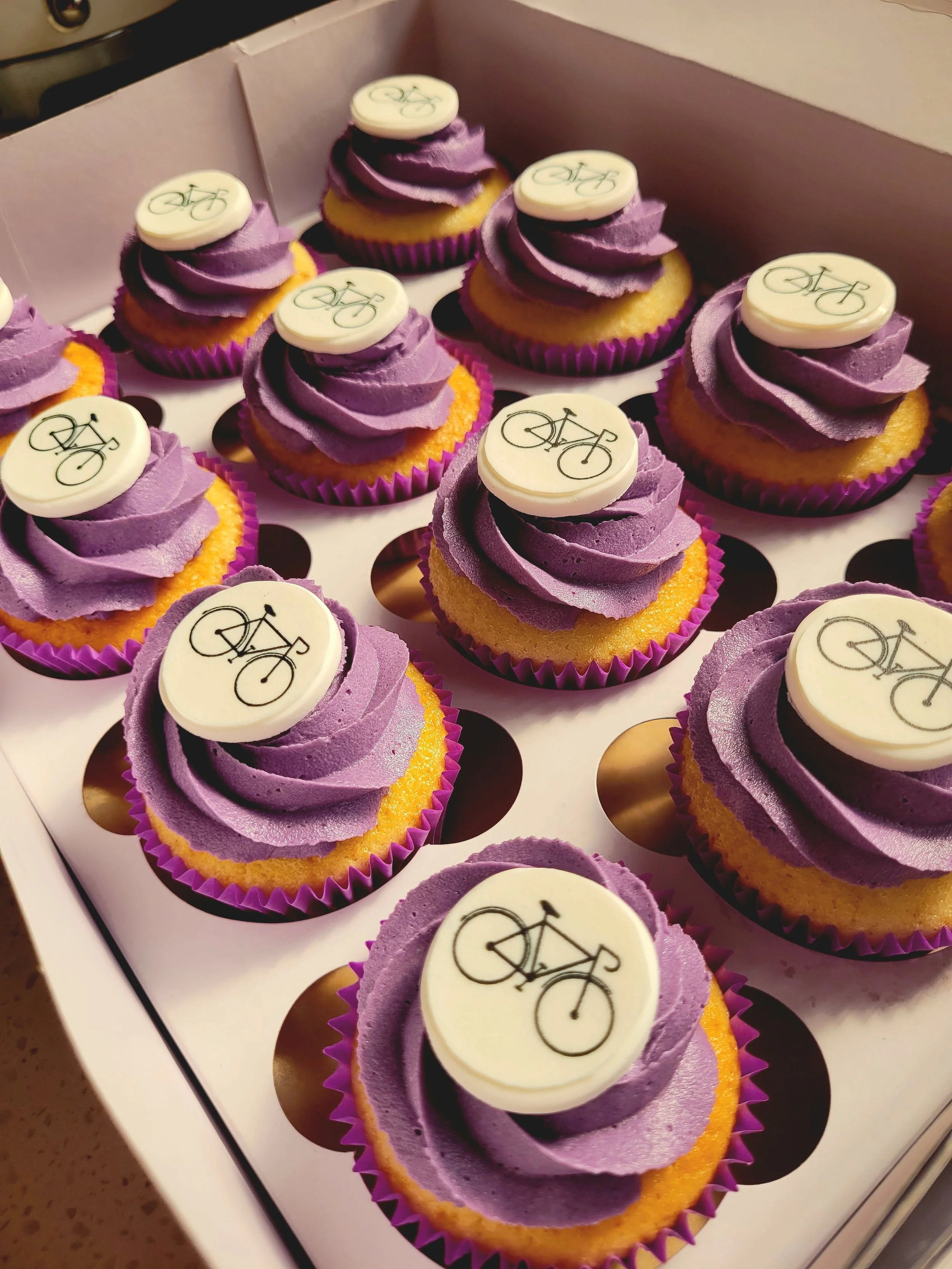 Cupcakes with purple frosting and bicycle-themed edible toppers arranged in a box.