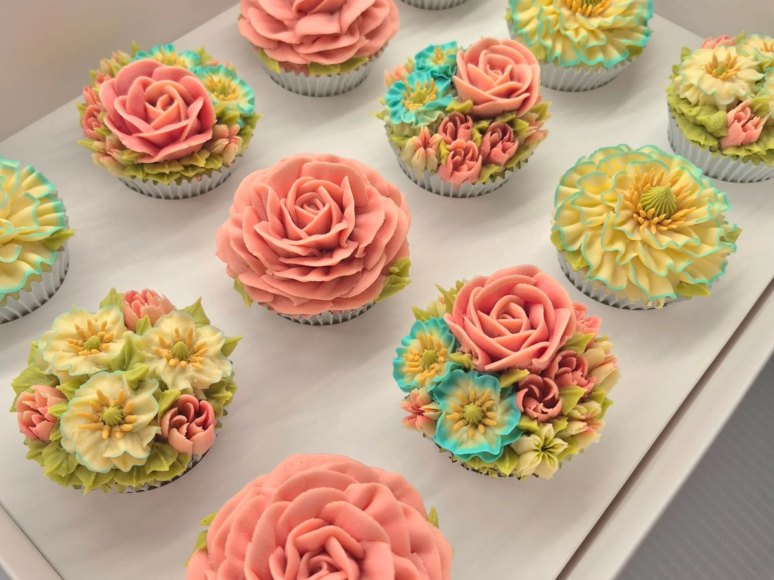 A collection of cupcakes decorated with colorful floral buttercream designs including pink roses, cream flowers with blue accents, and green leaves