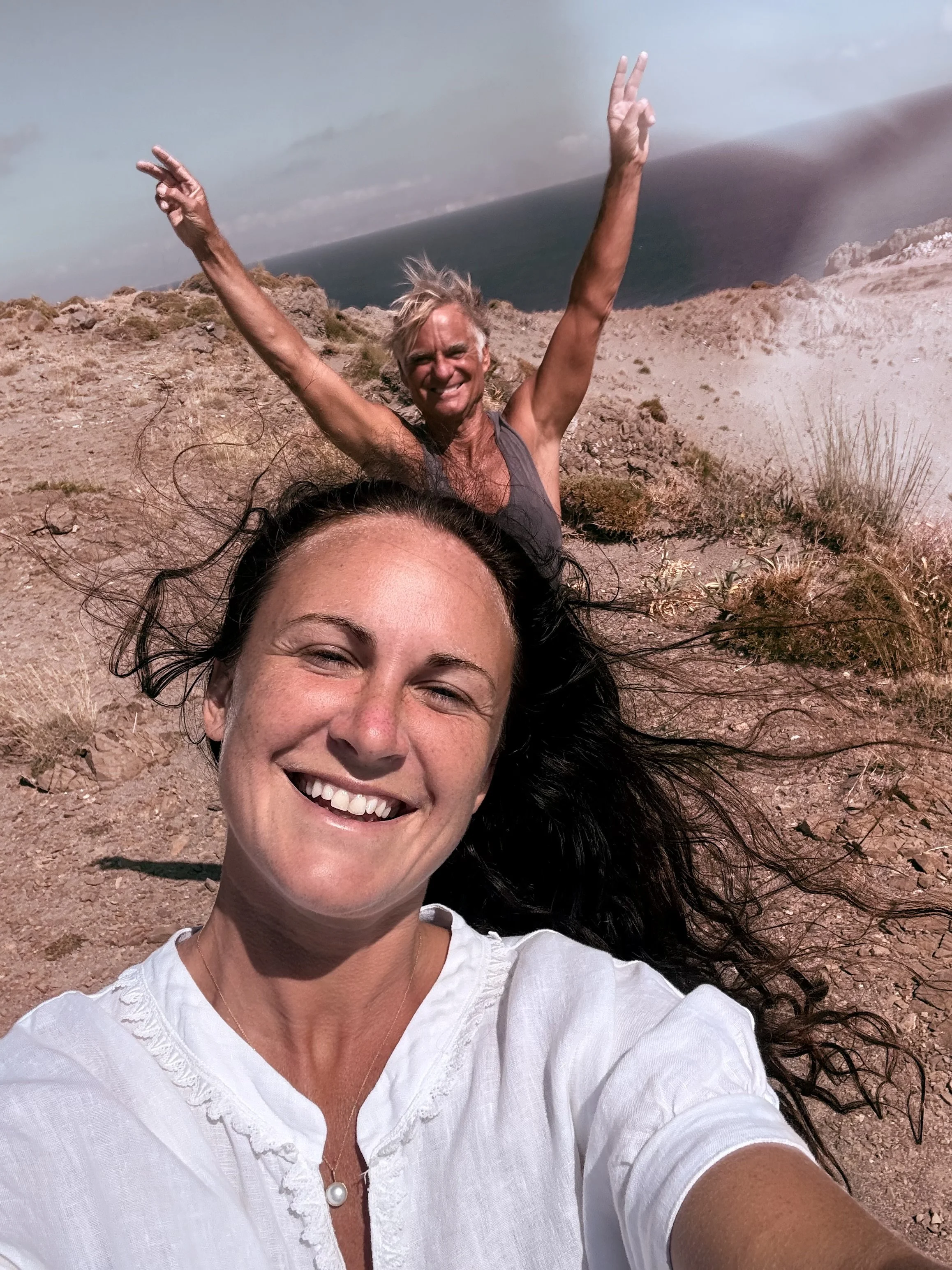 A woman with dark hair taking a selfie on a rocky, desert-like landscape with a man in the background, arms raised, smiling, and wind blowing.