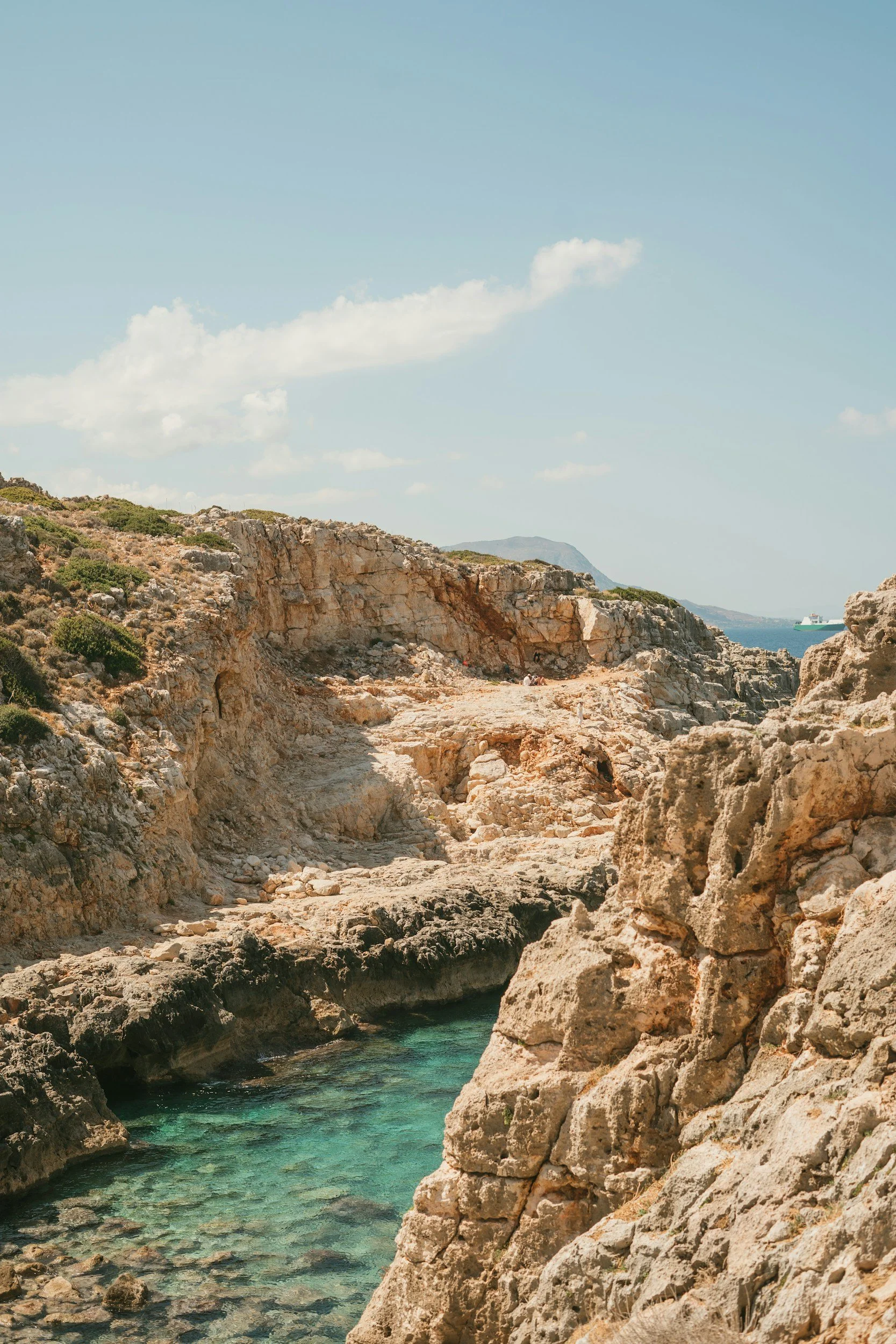 Coastal cliffs with rocky formations and clear turquoise water below, under a blue sky with a few clouds, and a ship in the distance on the water.