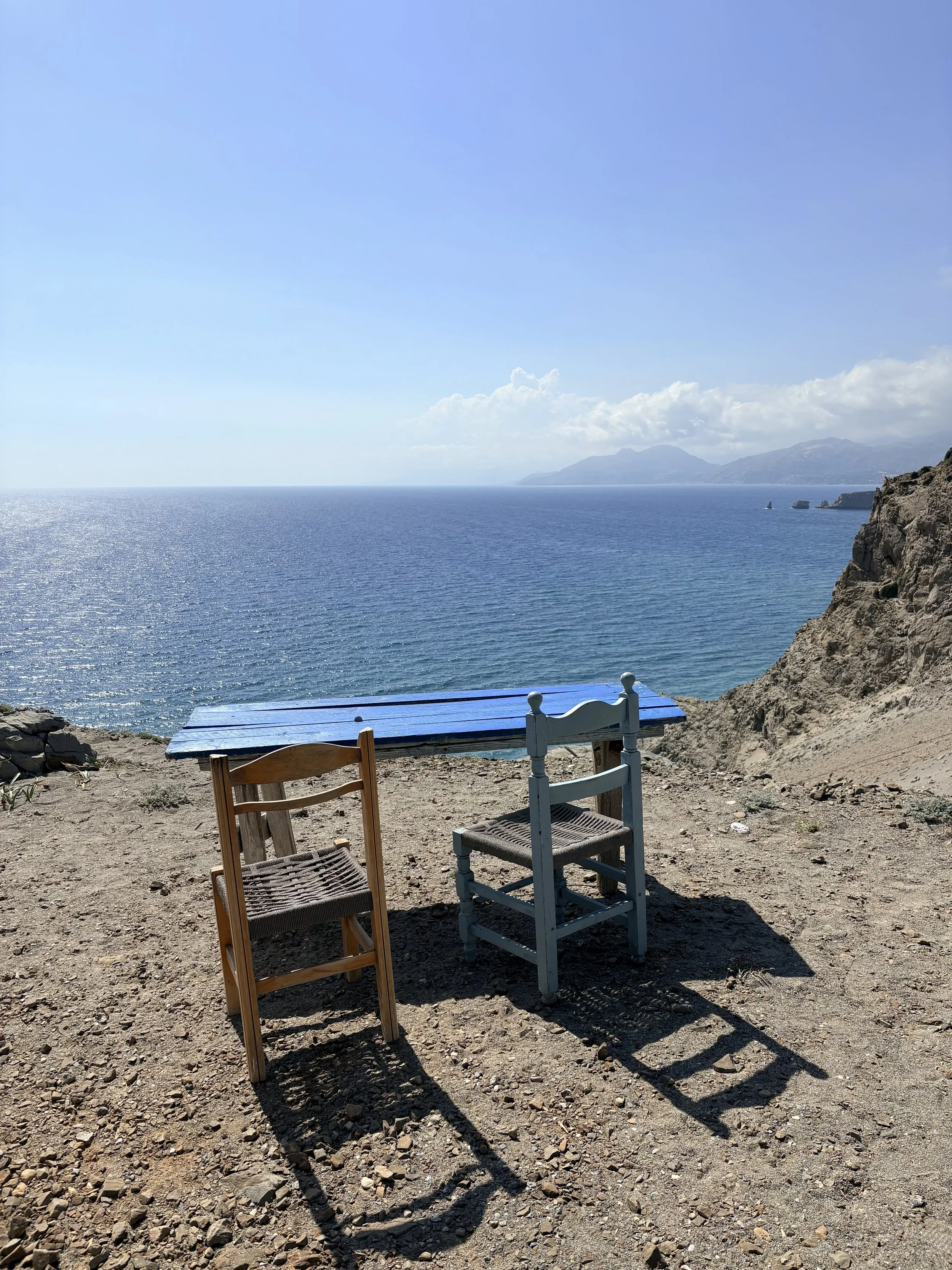 Seaside view with three chairs and a blue wooden bench on rocky ground facing ocean and distant mountains, partly sunny sky.