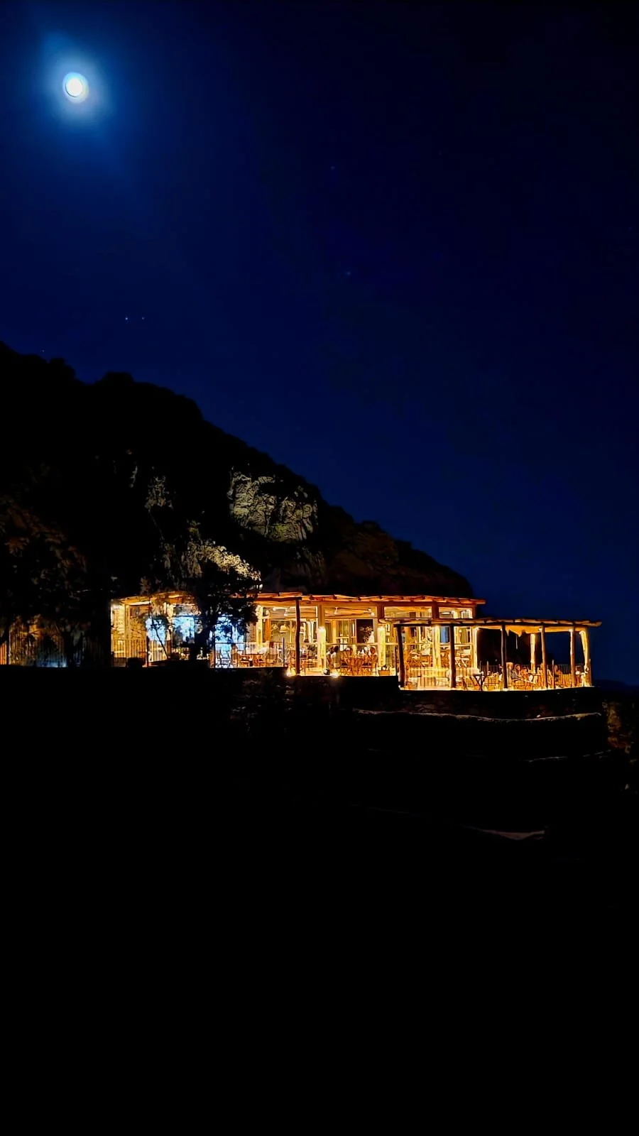 Night scene of a lit restaurant or cafe on a hillside overlooking the ocean, with a bright moon and a few stars in the dark sky.